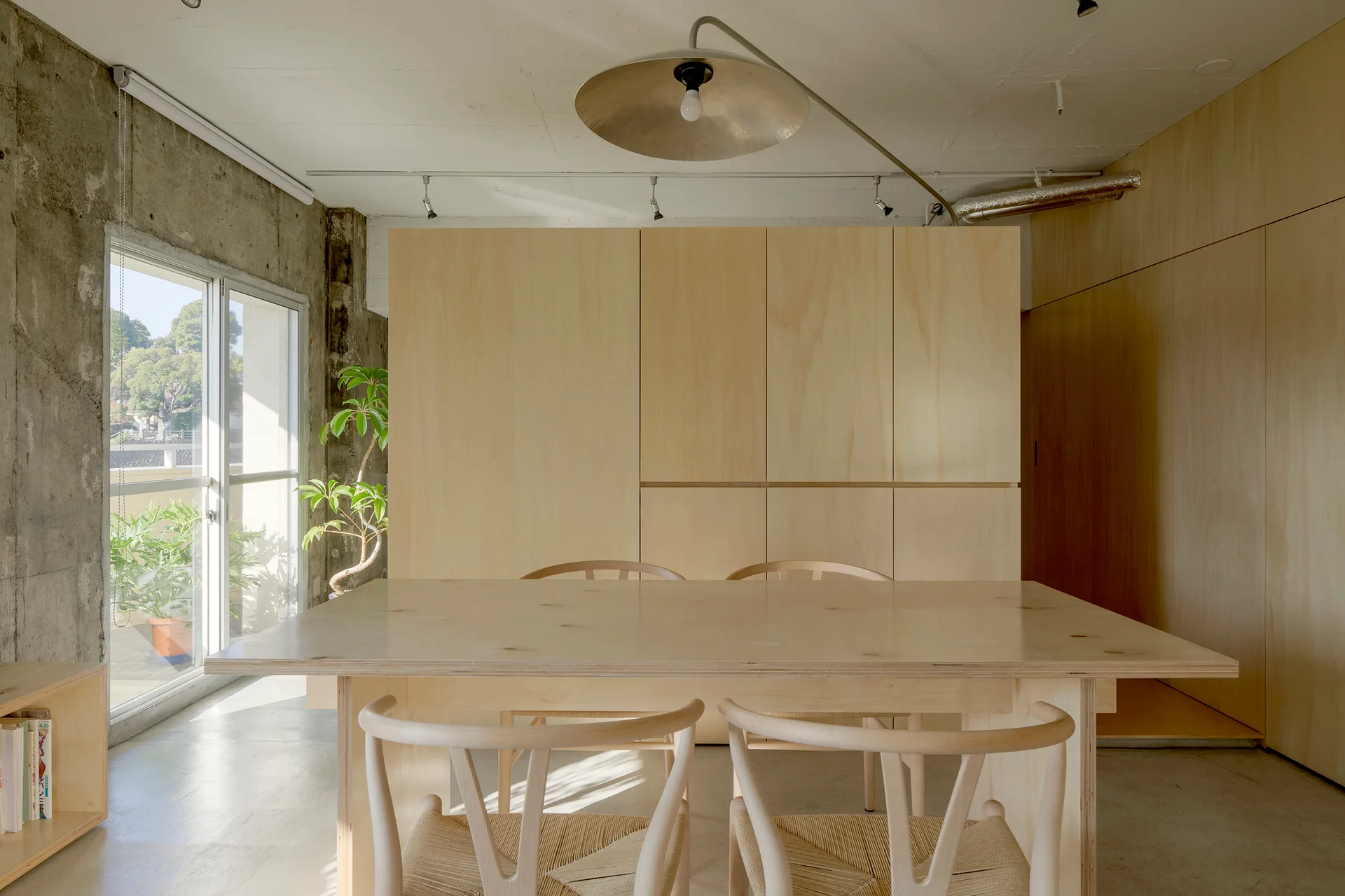A minimalist dining room features a light wood table and woven-seat chairs set against a large plywood wall and raw concrete partition.