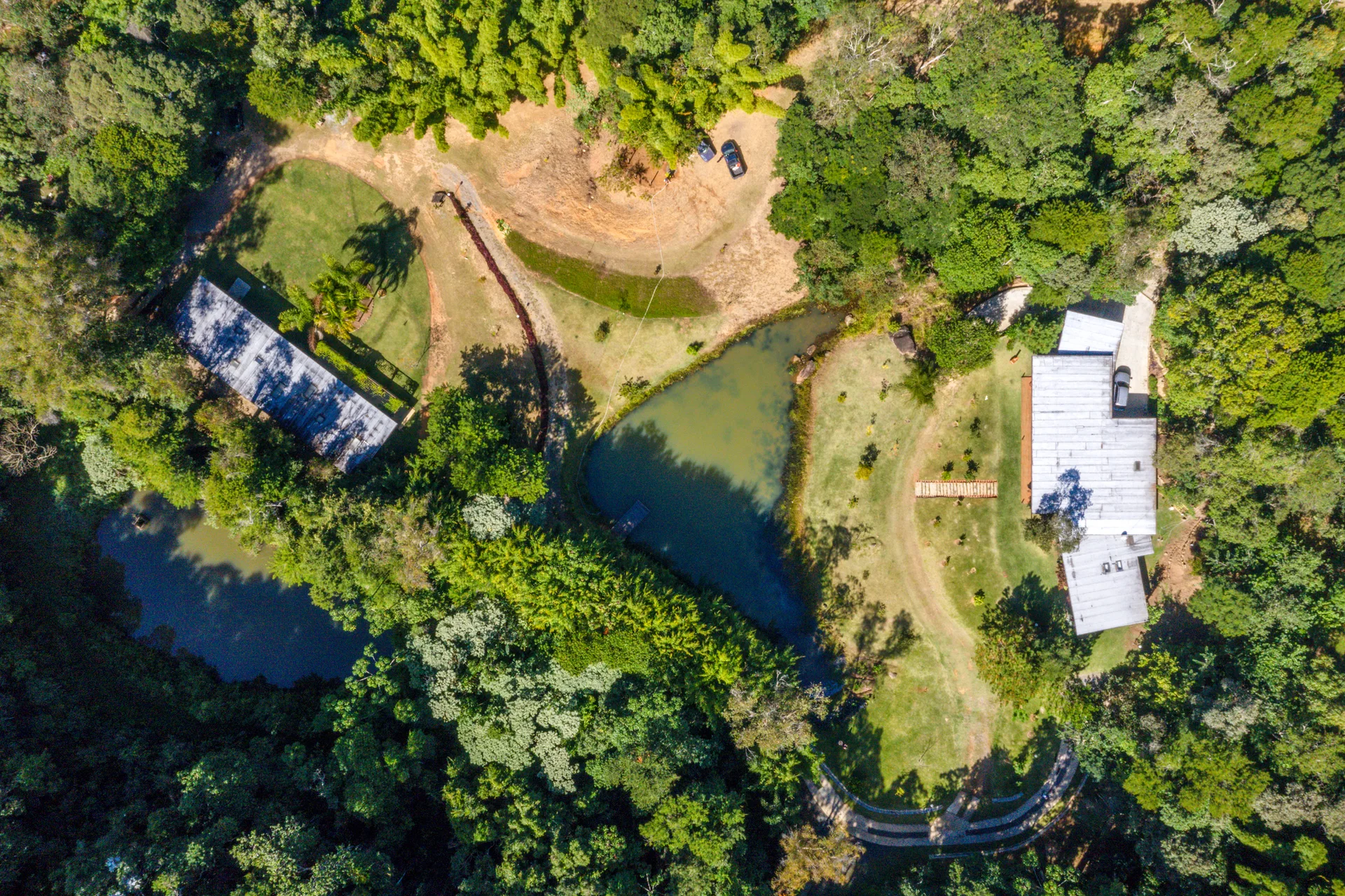 An aerial perspective of a secluded green property showing two houses with silver roofs, manicured lawns, and two ponds surrounded by dense trees.