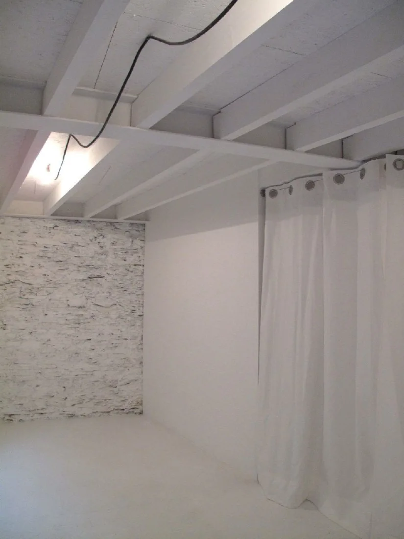 A minimalist white room features exposed wooden ceiling beams, a textured white brick wall, and a hanging white curtain.
