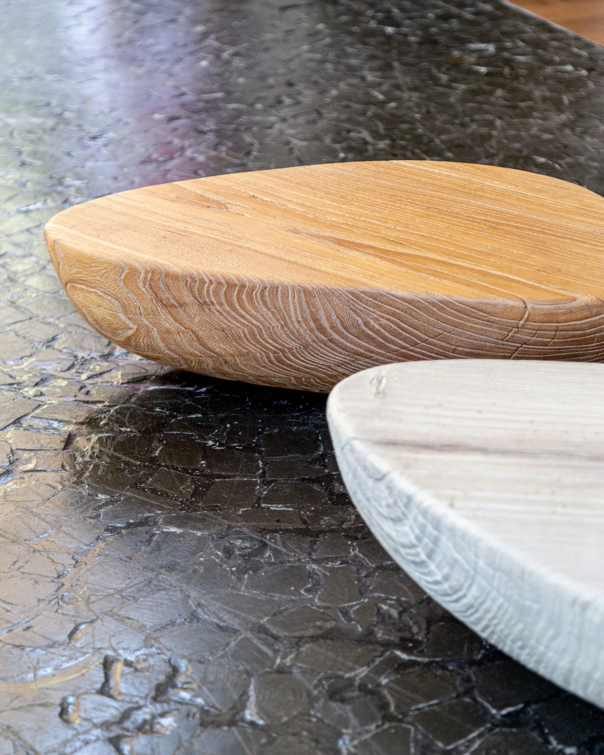 Two teardrop-shaped wooden sculptures, one light grey and one warm brown, rest on a textured, dark grey surface with a cracked pattern.
