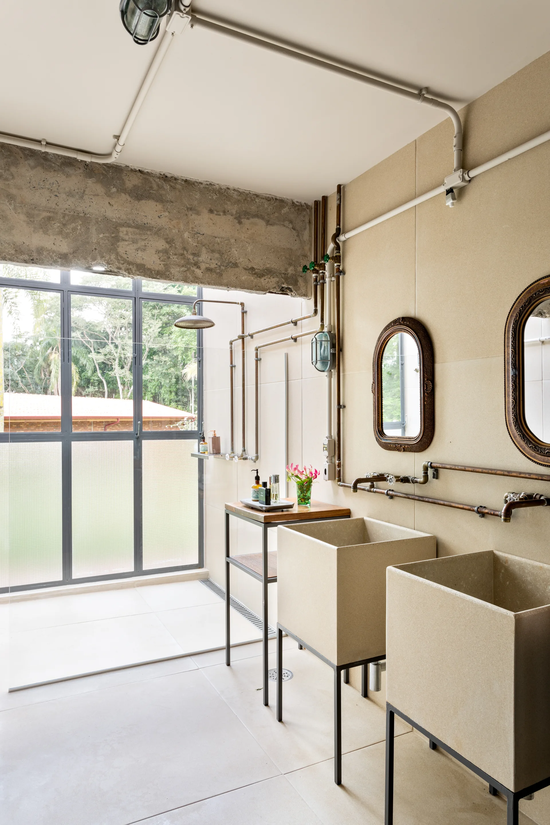 An industrial bathroom features two rectangular beige stone basins on metal frames, exposed copper piping, and ornate oval mirrors against a tiled wall.