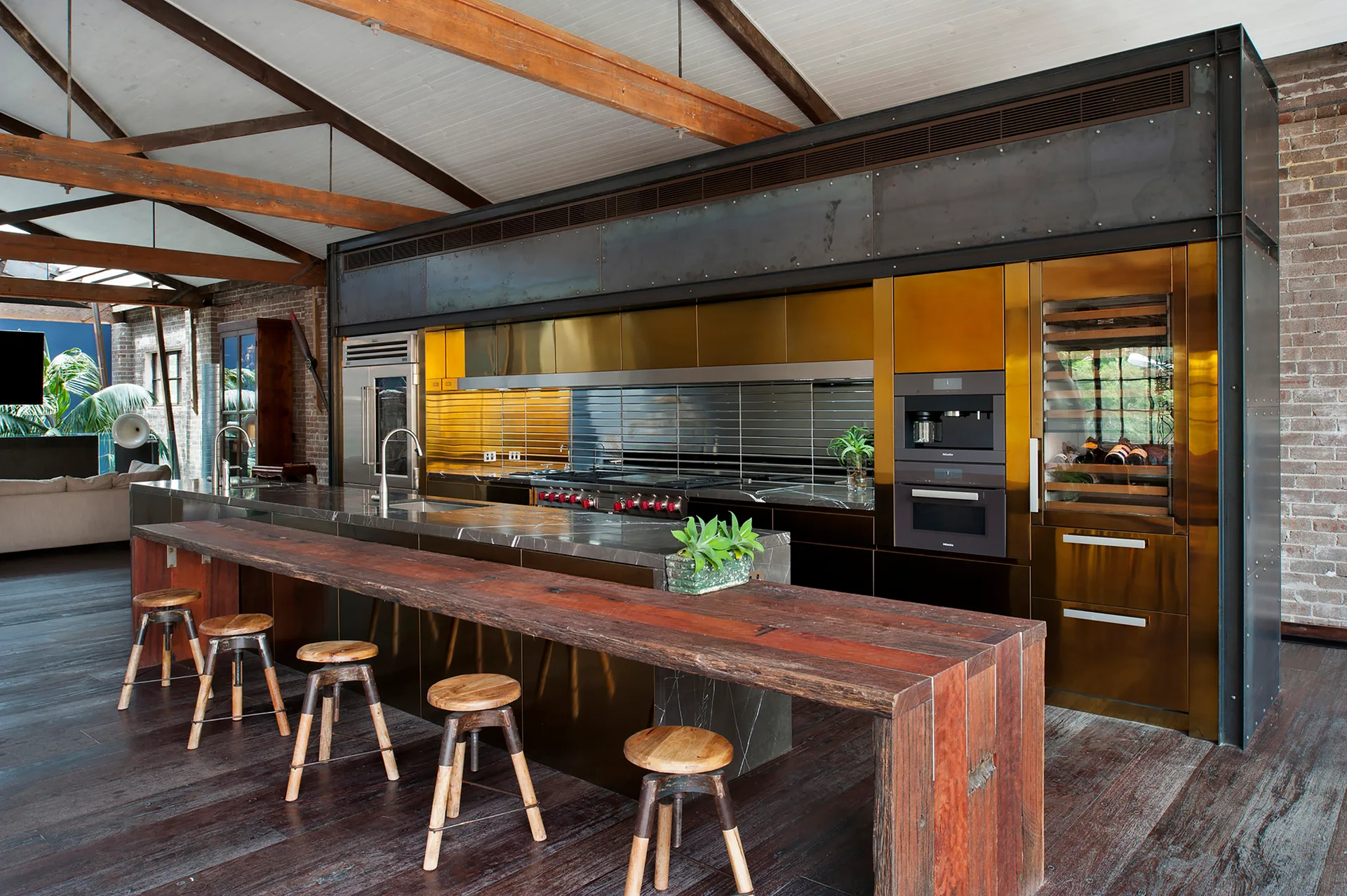 An industrial loft kitchen featuring brass-toned cabinetry, a dark stone island with a reclaimed wood breakfast bar, and exposed ceiling beams.