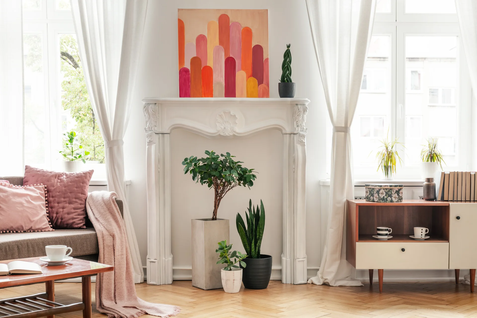 A light-filled living room featuring an ornate white fireplace mantel styled with plants inside and colorful abstract art on top.