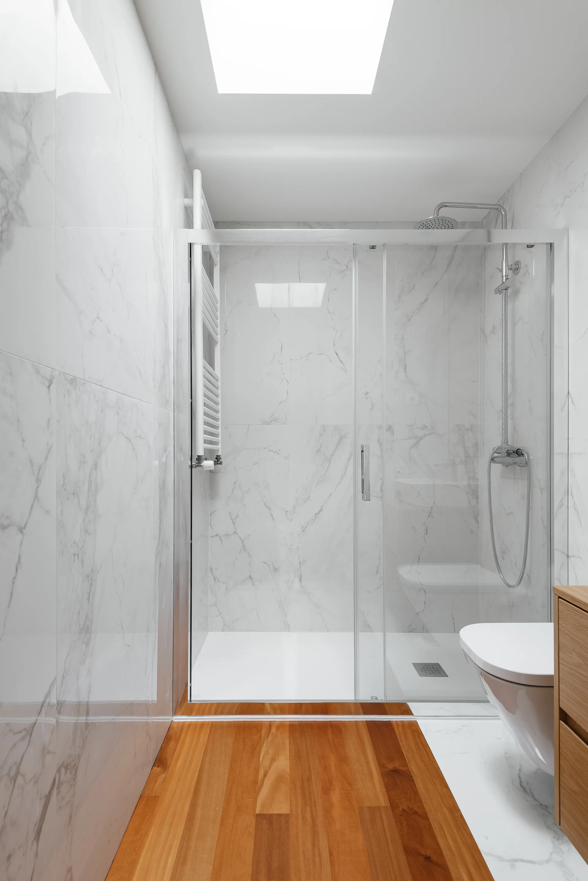 A modern bathroom features large-format white marble wall tiles, a glass-enclosed walk-in shower, a wall-mounted toilet, and a warm wood plank floor.