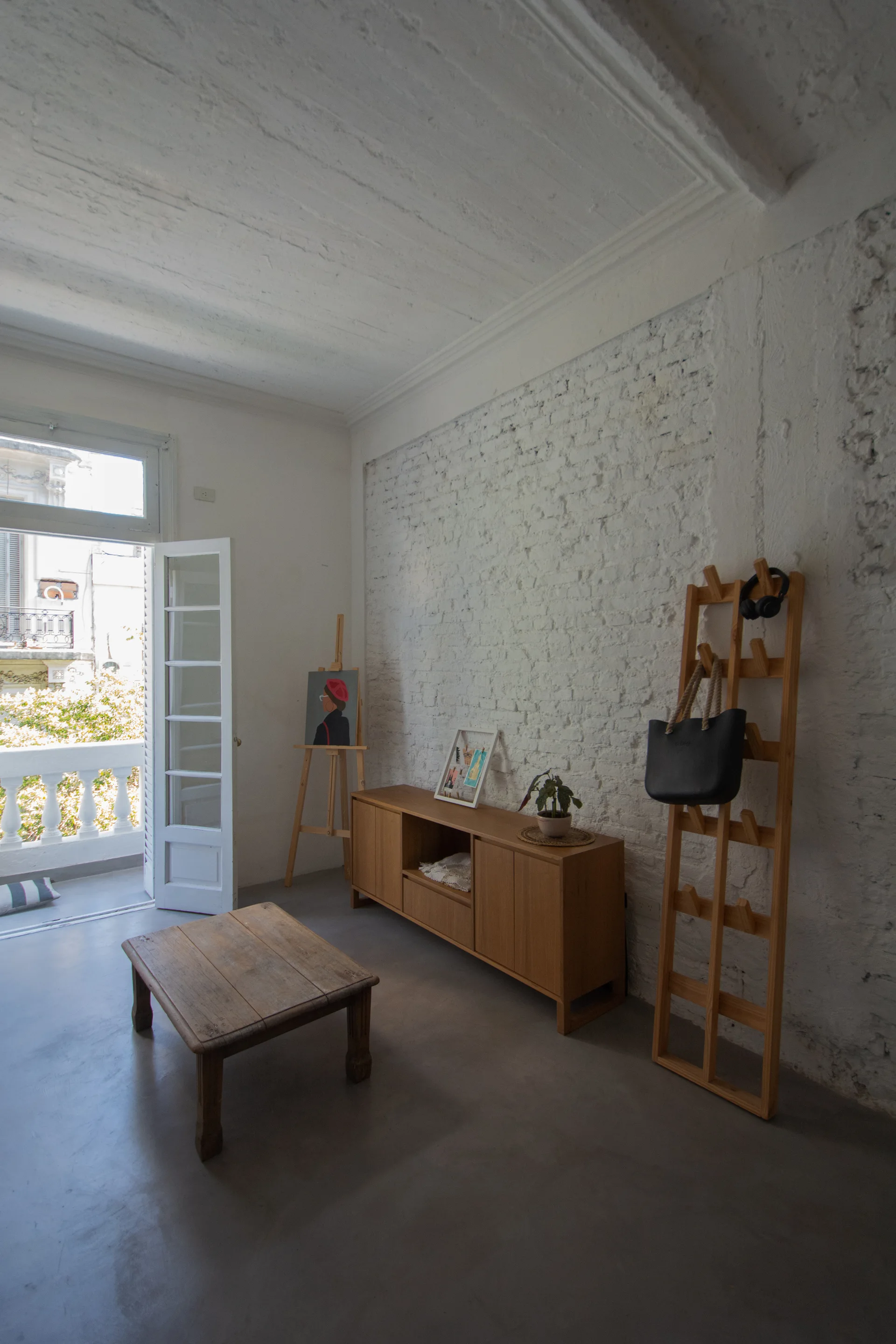 A minimalist living room features a wooden coffee table and sideboard against a white exposed brick wall next to an open balcony door.