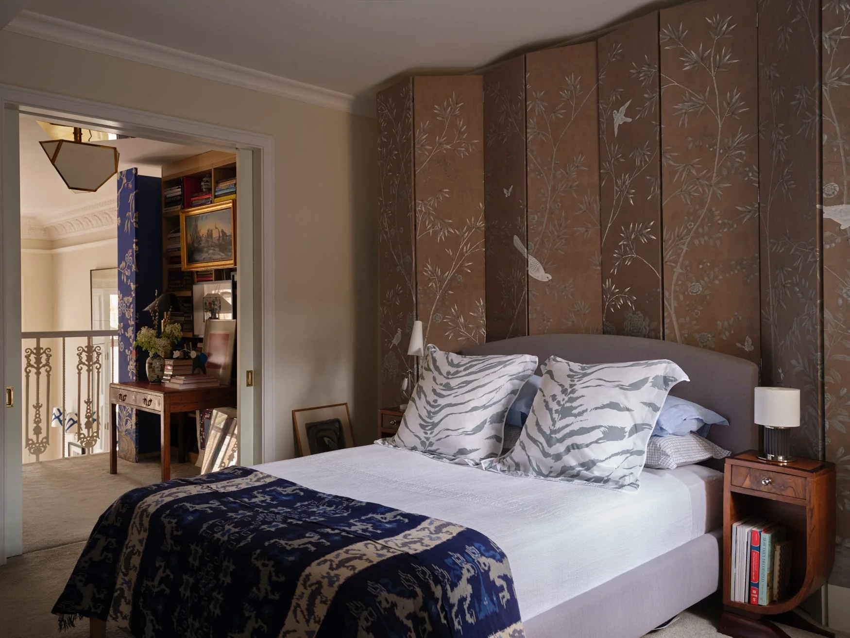 A bedroom featuring a grey upholstered headboard, a brown botanical patterned screen wall, tiger-print pillows, and a wooden nightstand.