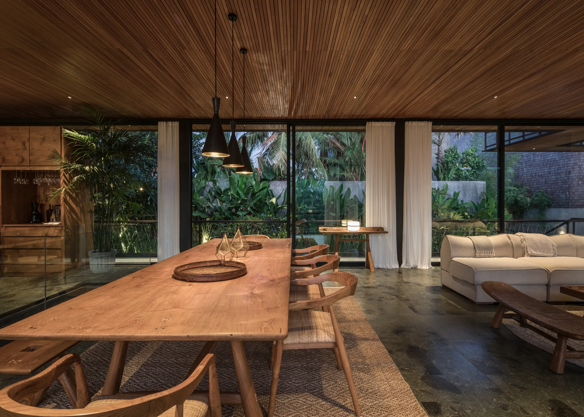 An open-concept dining room features a long wooden table, matching chairs with woven seats, a wood-slatted ceiling, and floor-to-ceiling glass walls.