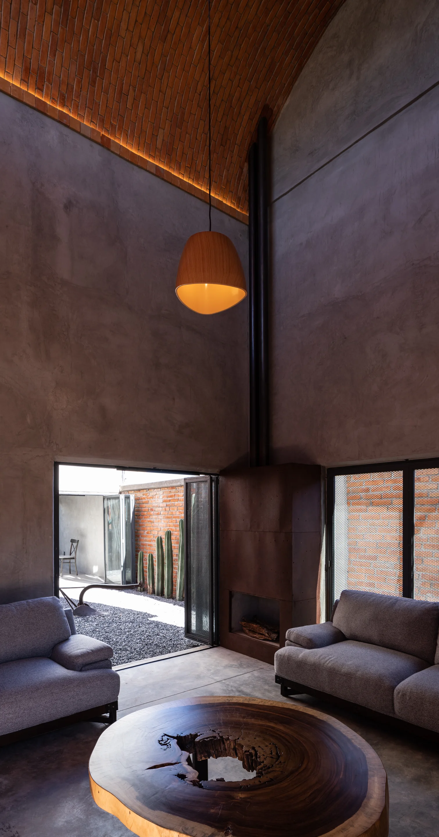 A modern living room with tall concrete walls, a vaulted brick ceiling, a rustic metal fireplace, and a live-edge wood coffee table.