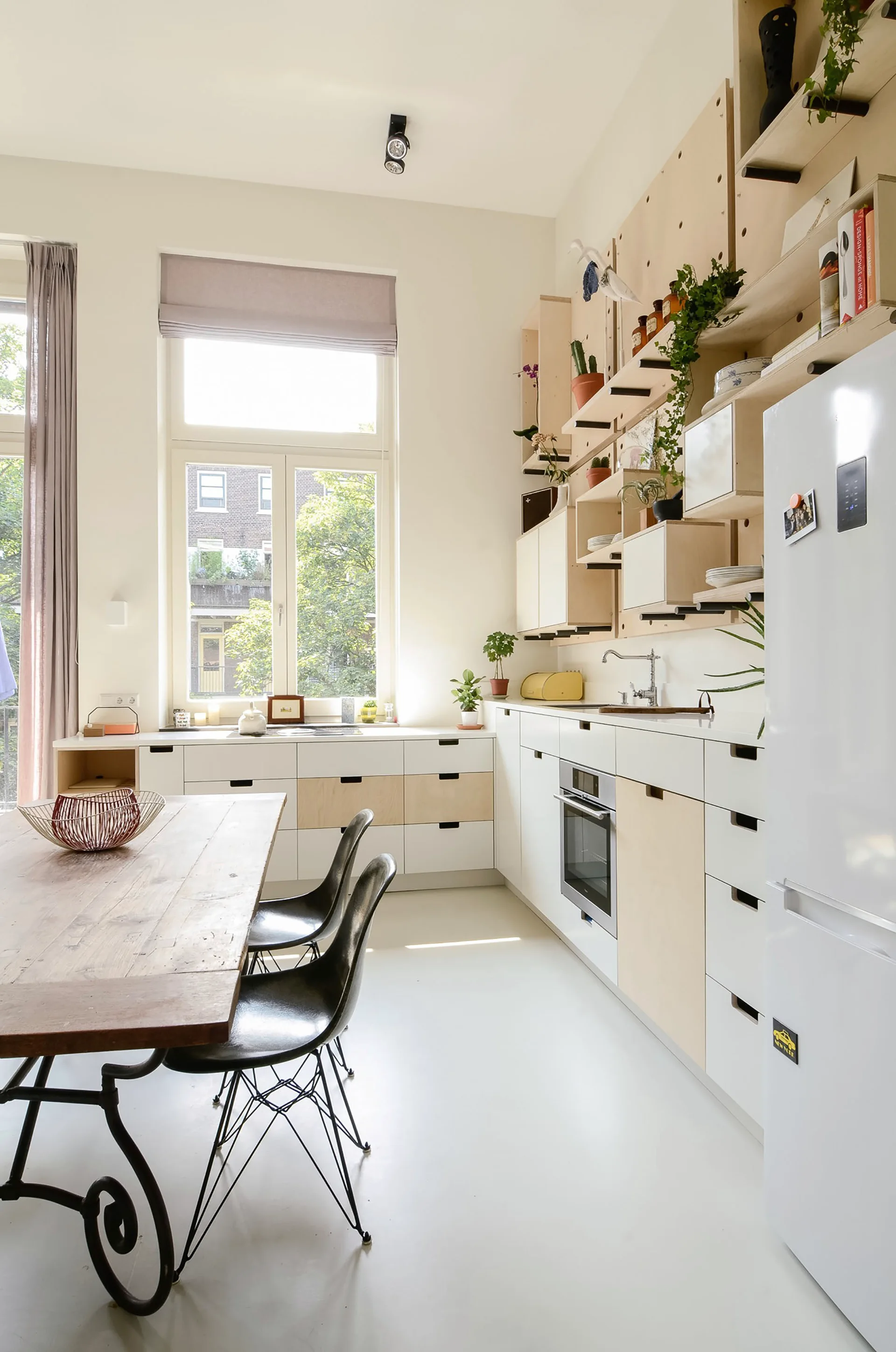 A bright kitchen features white and light wood cabinetry, a wooden dining table with black chairs, and a large plywood pegboard storage system.