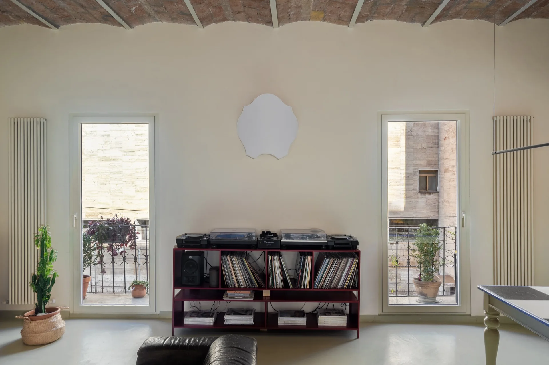 A minimalist living room features a vaulted brick ceiling, two balcony doors, and a deep red console holding record players and vinyl records.