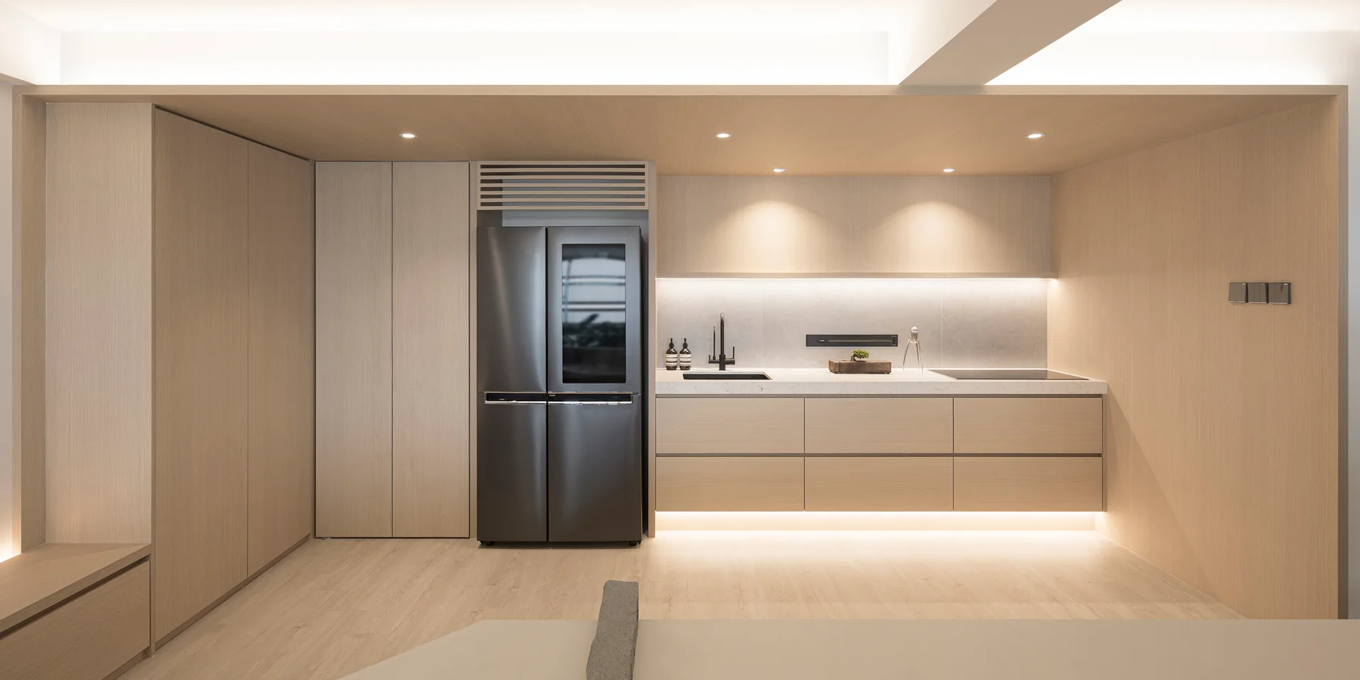 A modern minimalist kitchen with handleless light wood cabinetry, an integrated black refrigerator, and warm under-cabinet lighting.