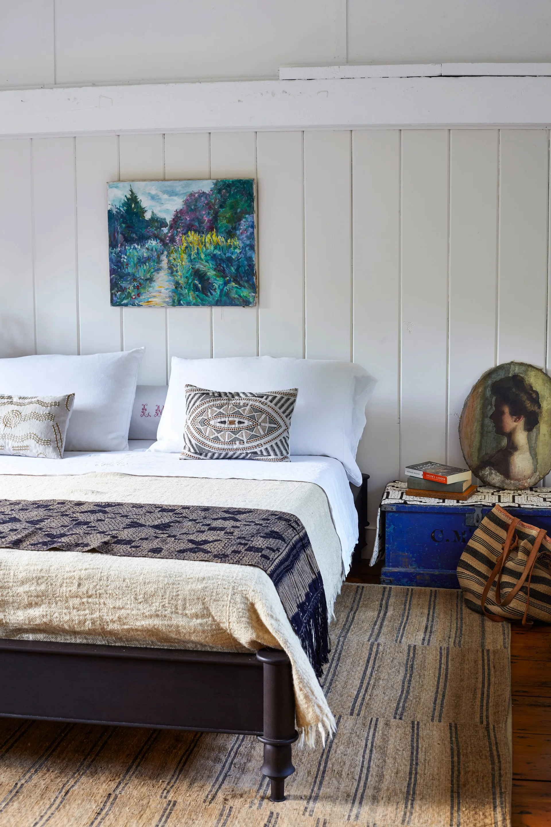 A bed with patterned textiles sits against a white vertical plank wall next to a blue wooden storage chest used as a nightstand with a portrait on top.