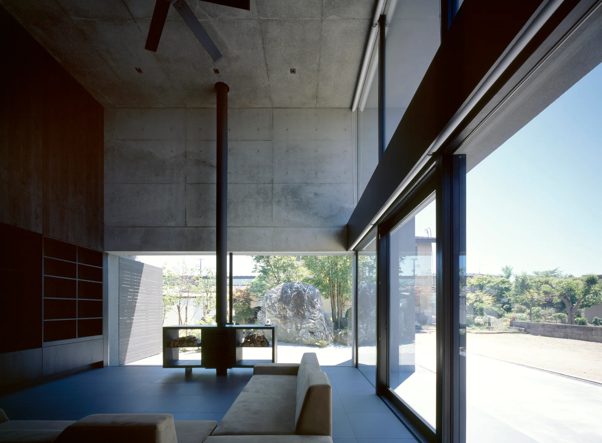 A minimalist living room features exposed concrete walls, a tan sectional sofa, and a black freestanding fireplace in front of large glass doors.