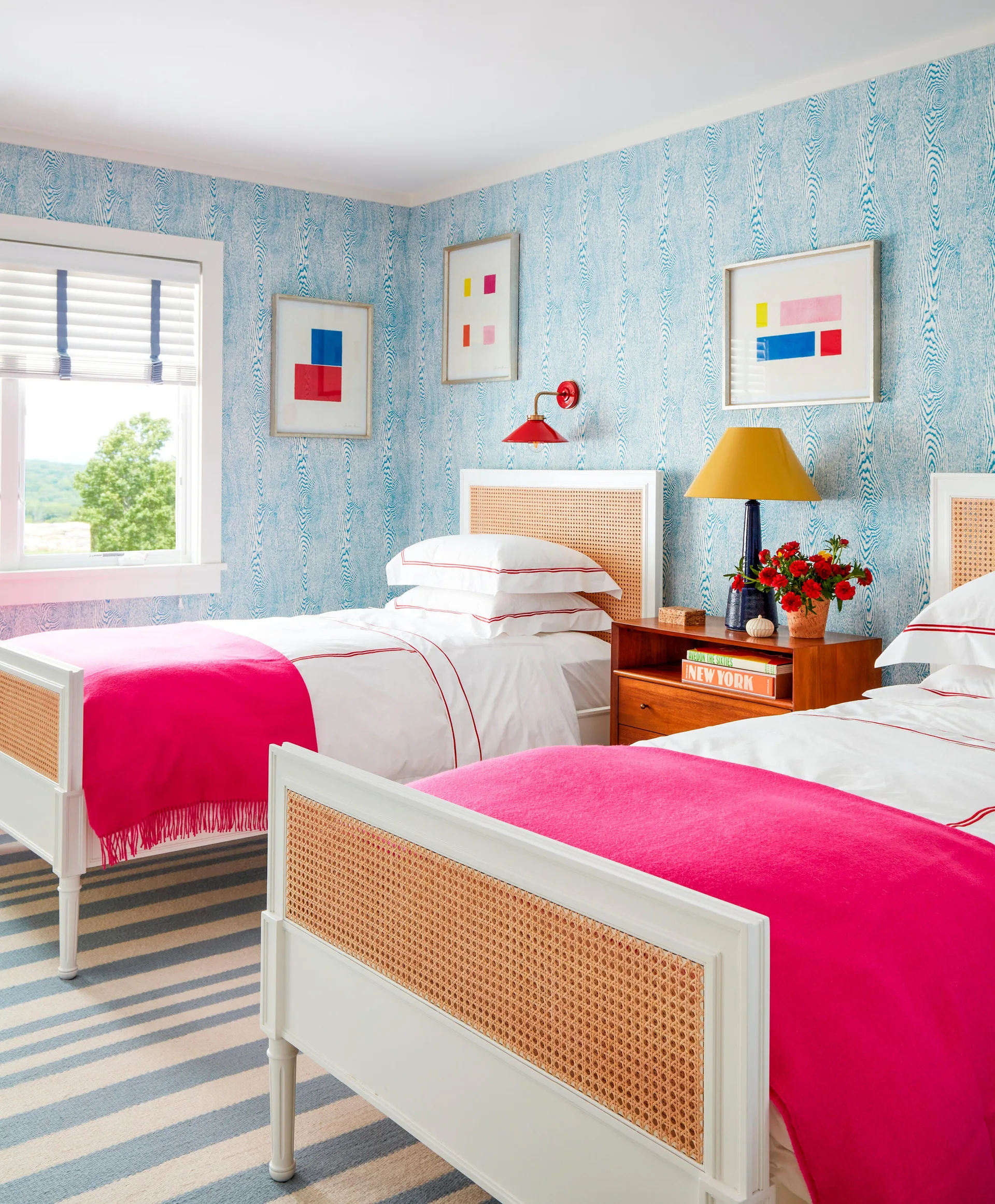 Two twin beds with white linens, hot pink throw blankets, and cane headboards sit against blue wood grain wallpaper with a striped rug below.