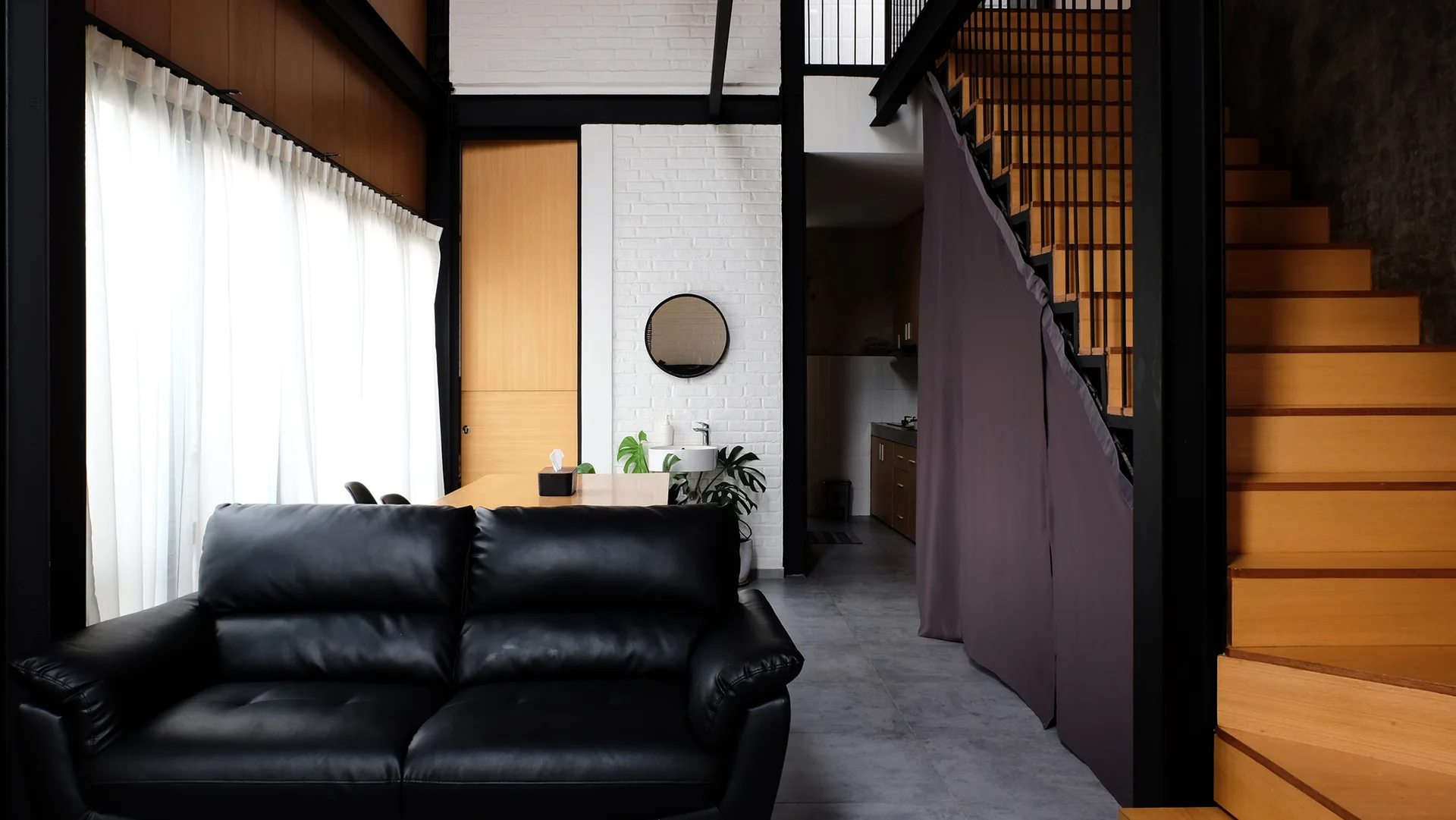 A modern living room features a black leather sofa, a wood-toned staircase with black metal railing, and an exposed white brick wall.