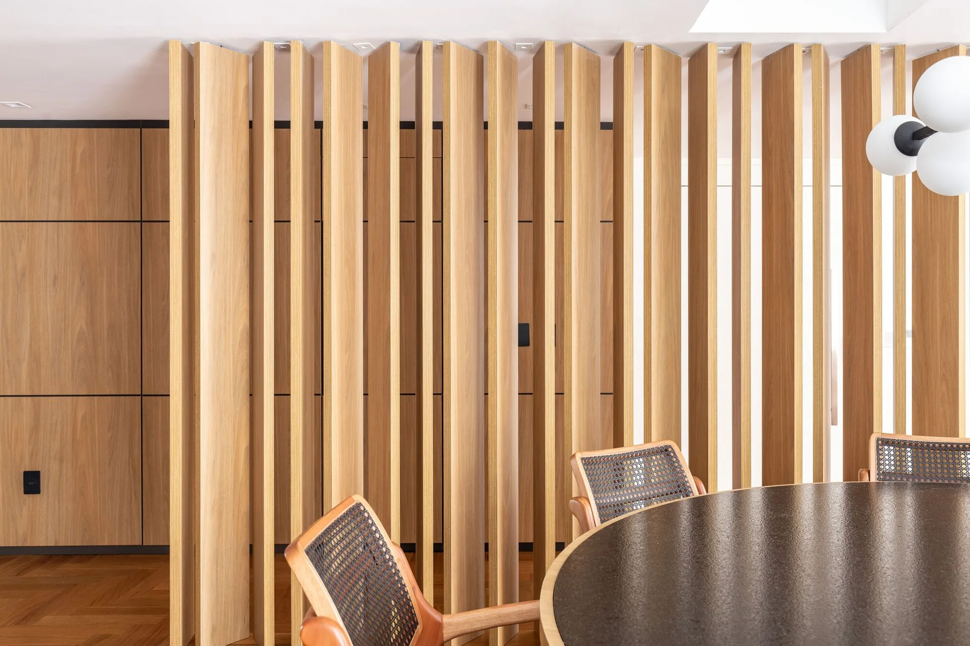 A dining area featuring a round dark brown table, cane-back chairs, and a vertical wooden slatted partition screen next to paneled walls.