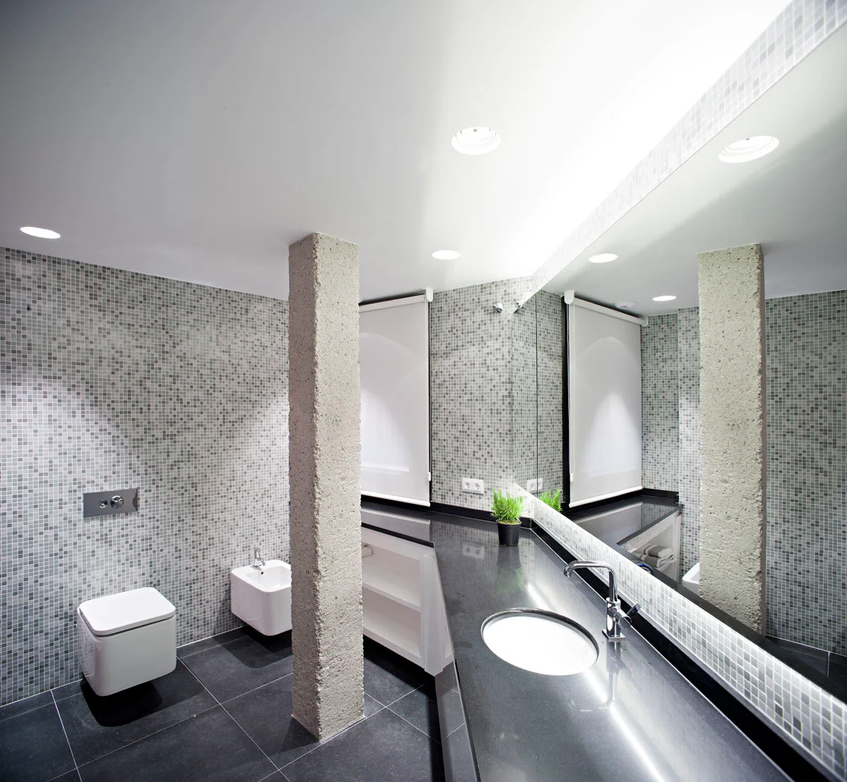 A modern bathroom features grey mosaic wall tiles, a black vanity countertop, a wall-mounted toilet, and a rough stone structural column.
