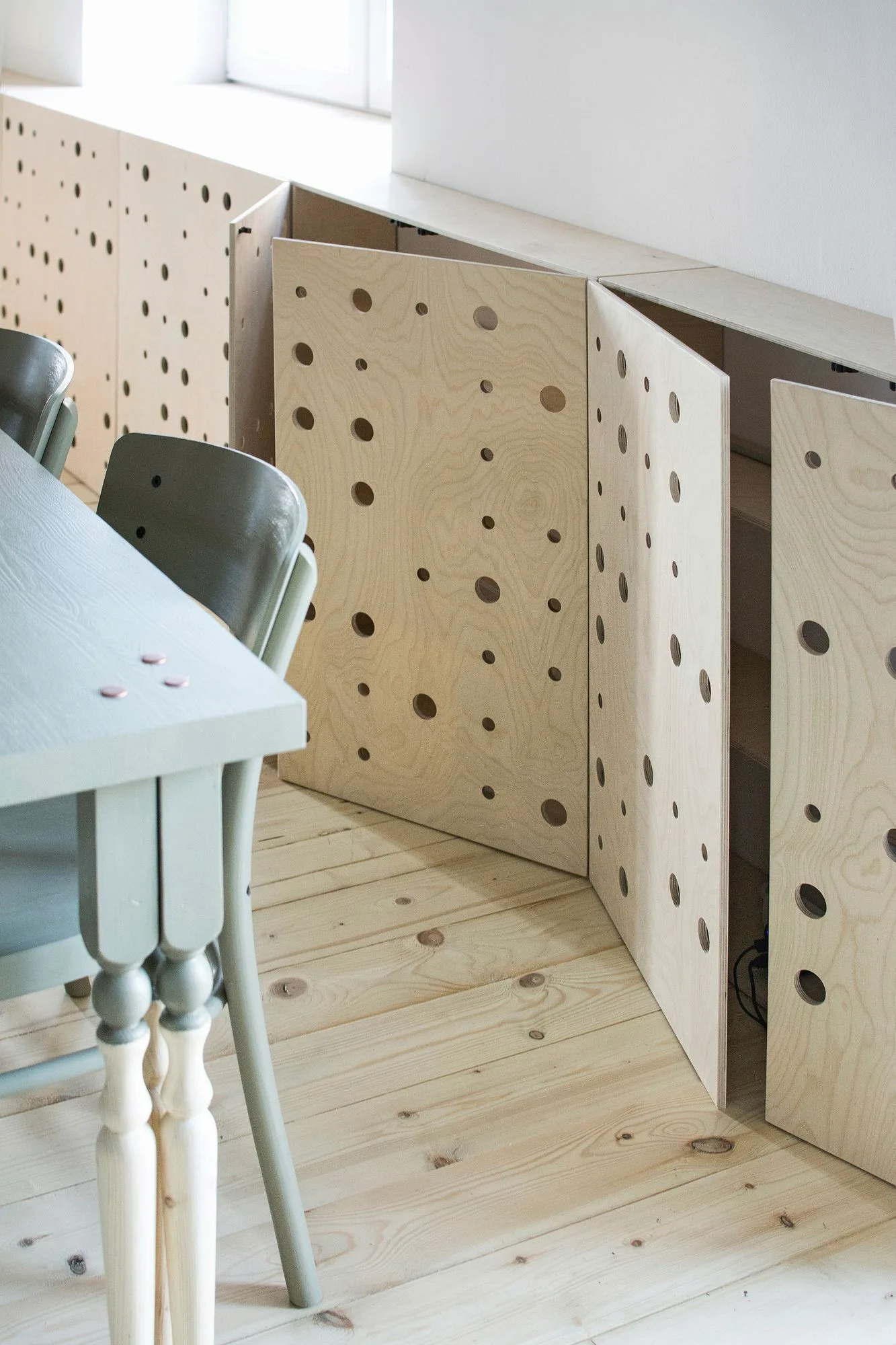 Custom plywood cabinets with circular perforated patterns sit on wood floors next to a sage green dining table and chairs.