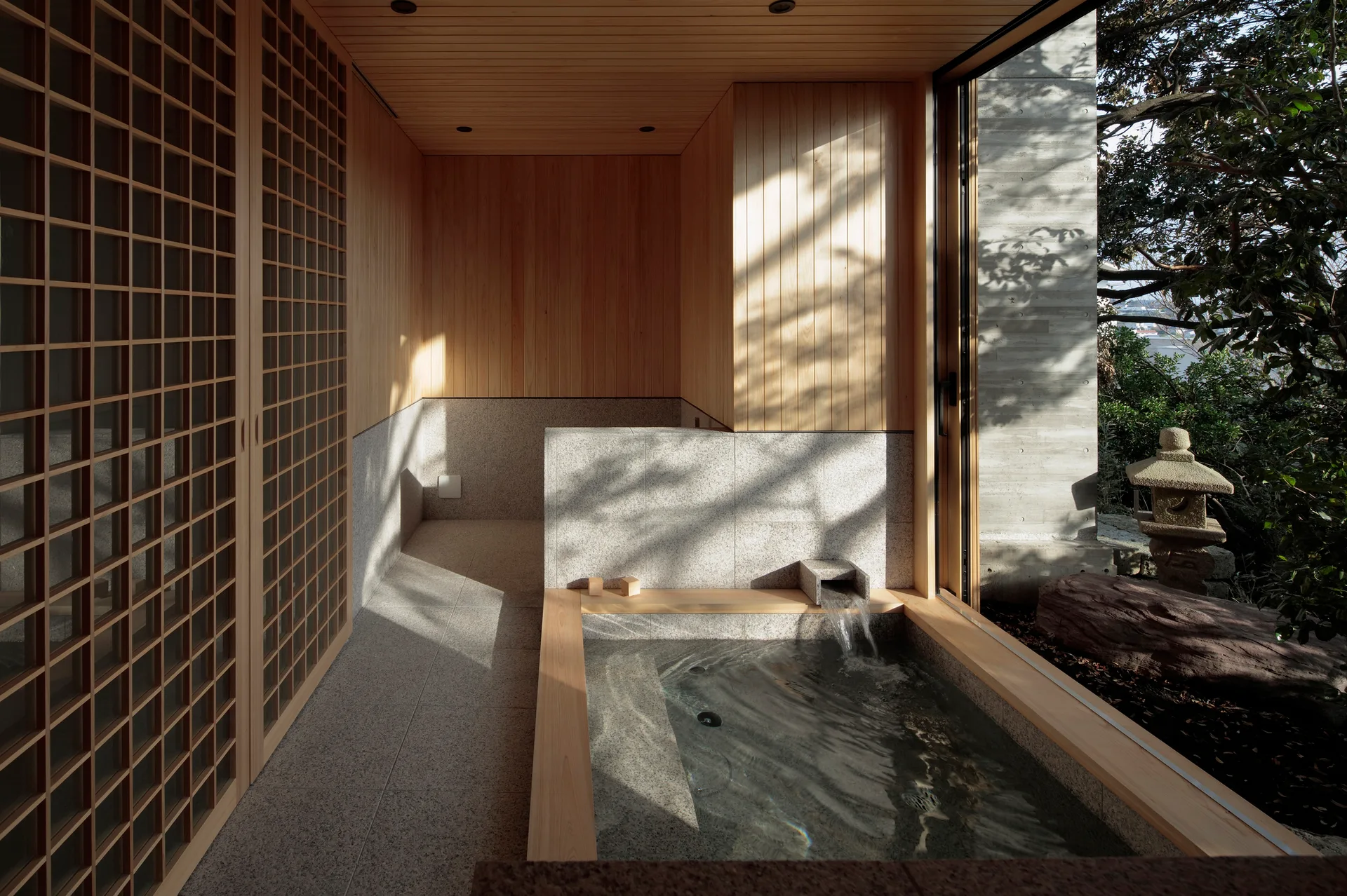 A minimalist Japanese-style spa room featuring a sunken stone soaking tub, wood-paneled walls, a sliding lattice screen, and a view of an outdoor garden.