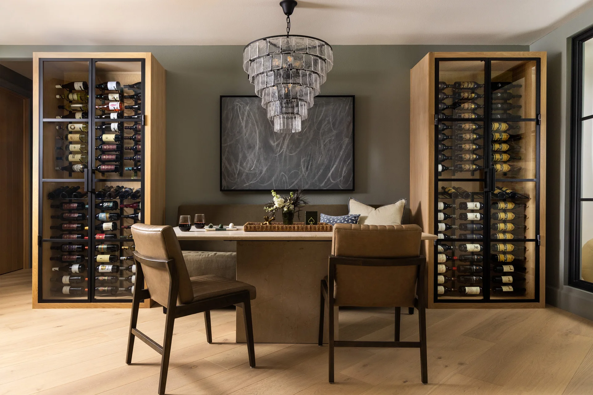 A dining area with a stone table, banquette, and tiered glass chandelier between two wood-framed wine cabinets on a herringbone floor.