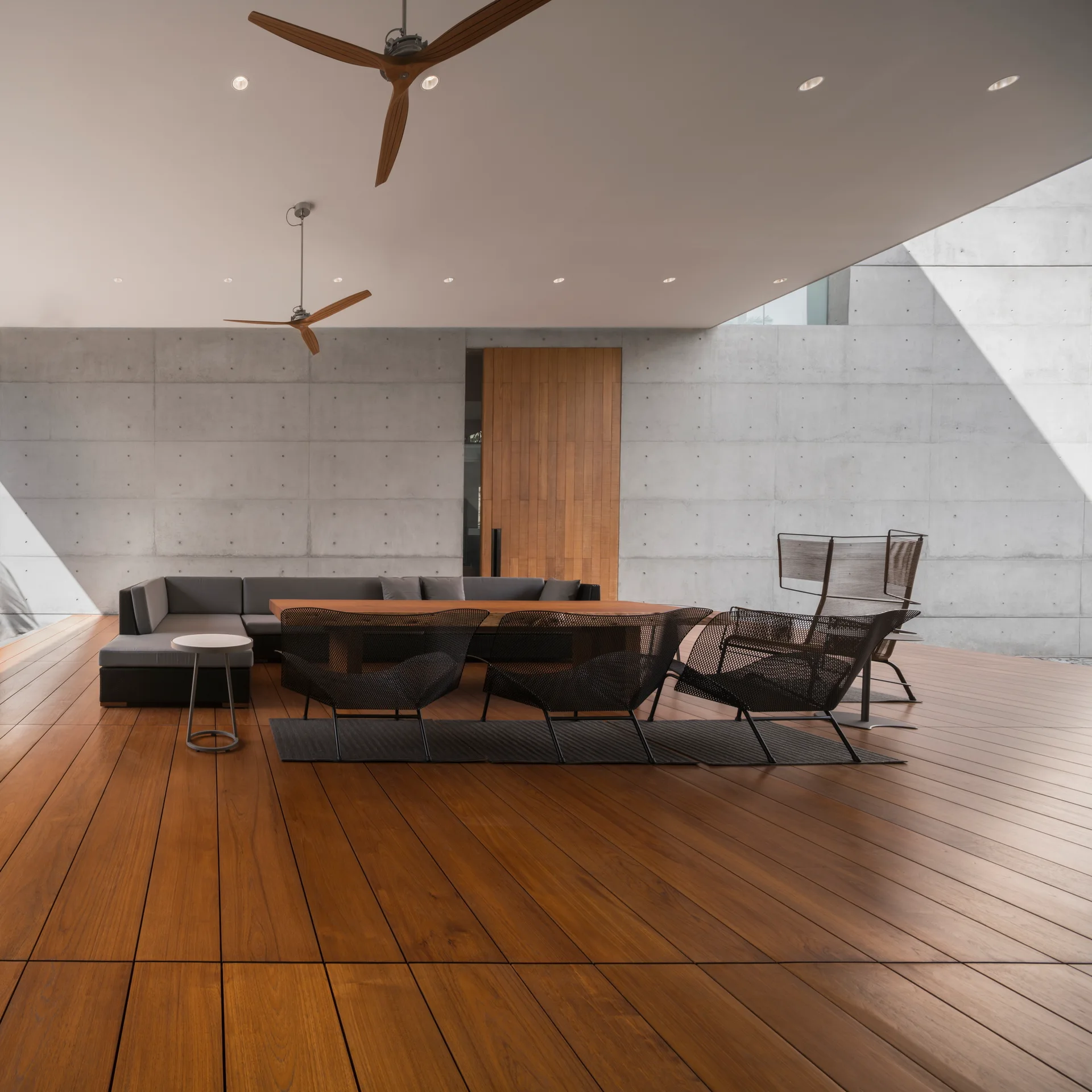 A covered outdoor patio features a warm wood deck, a grey concrete panel wall, and dark contemporary seating arranged under wooden ceiling fans.