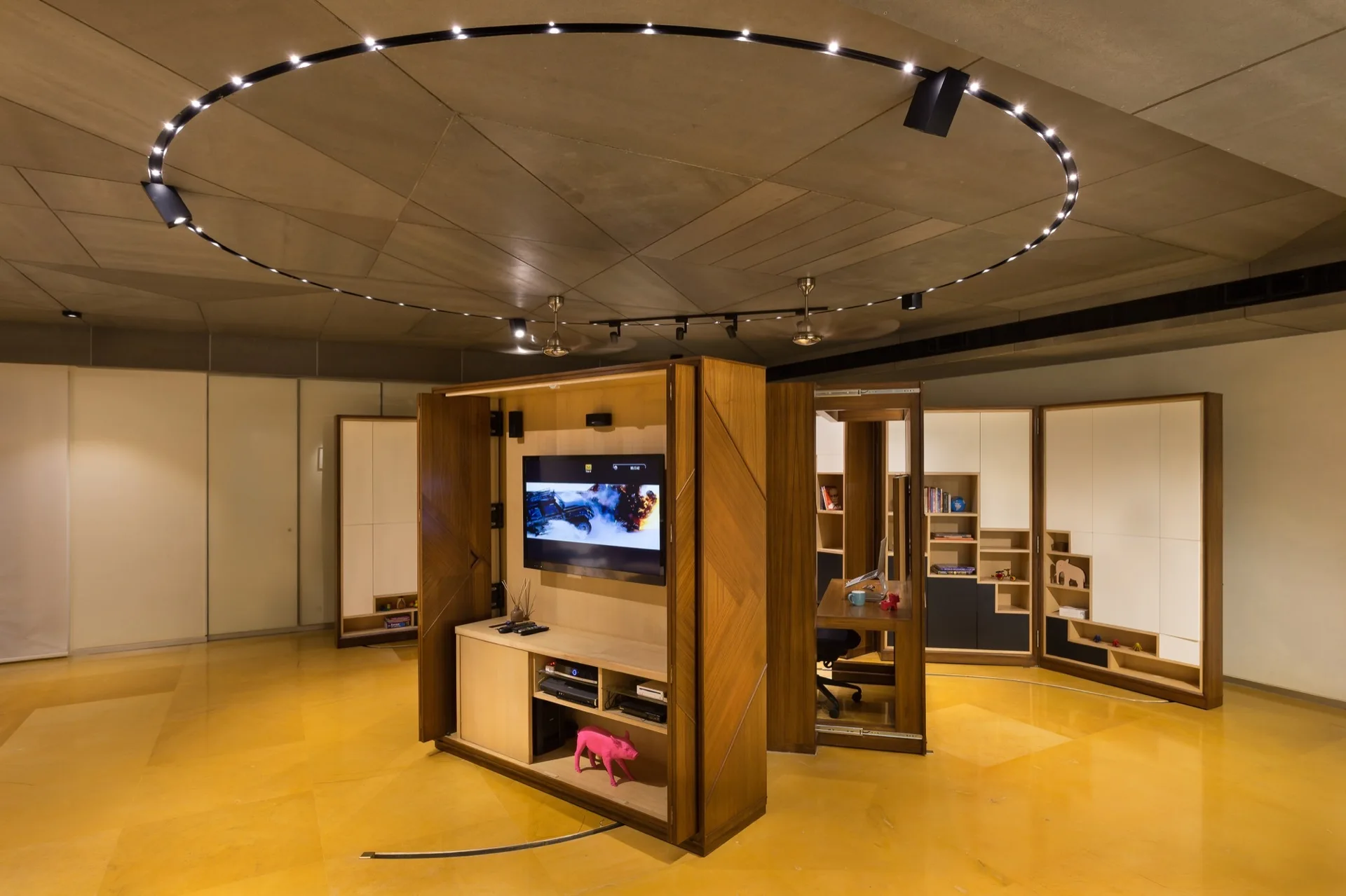 A central wooden modular cabinet serving as a media unit and desk stands on yellow stone floors under circular ceiling track lights.