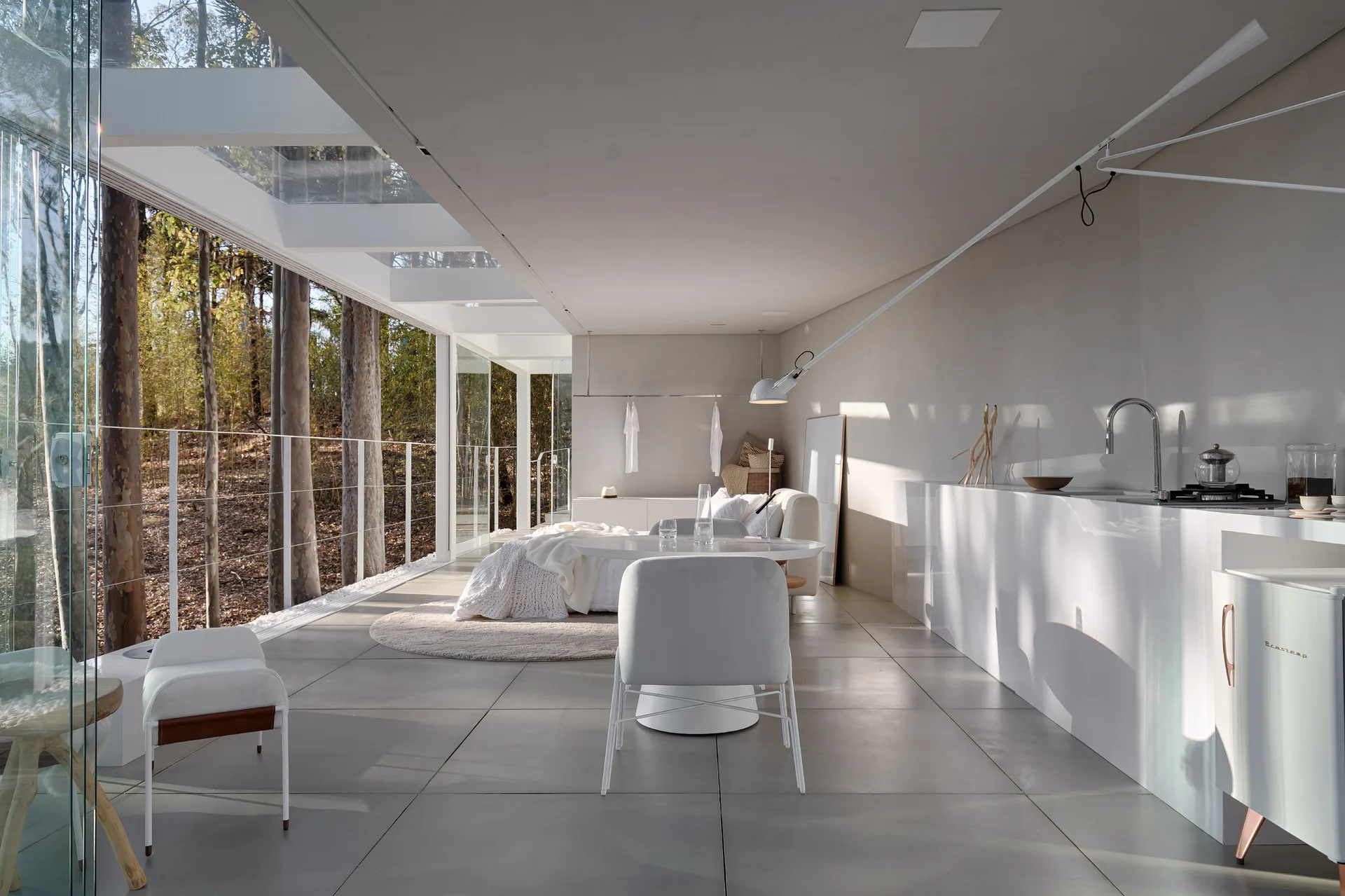 A minimalist white kitchen and dining area features large floor-to-ceiling glass walls showing an outdoor forest view.