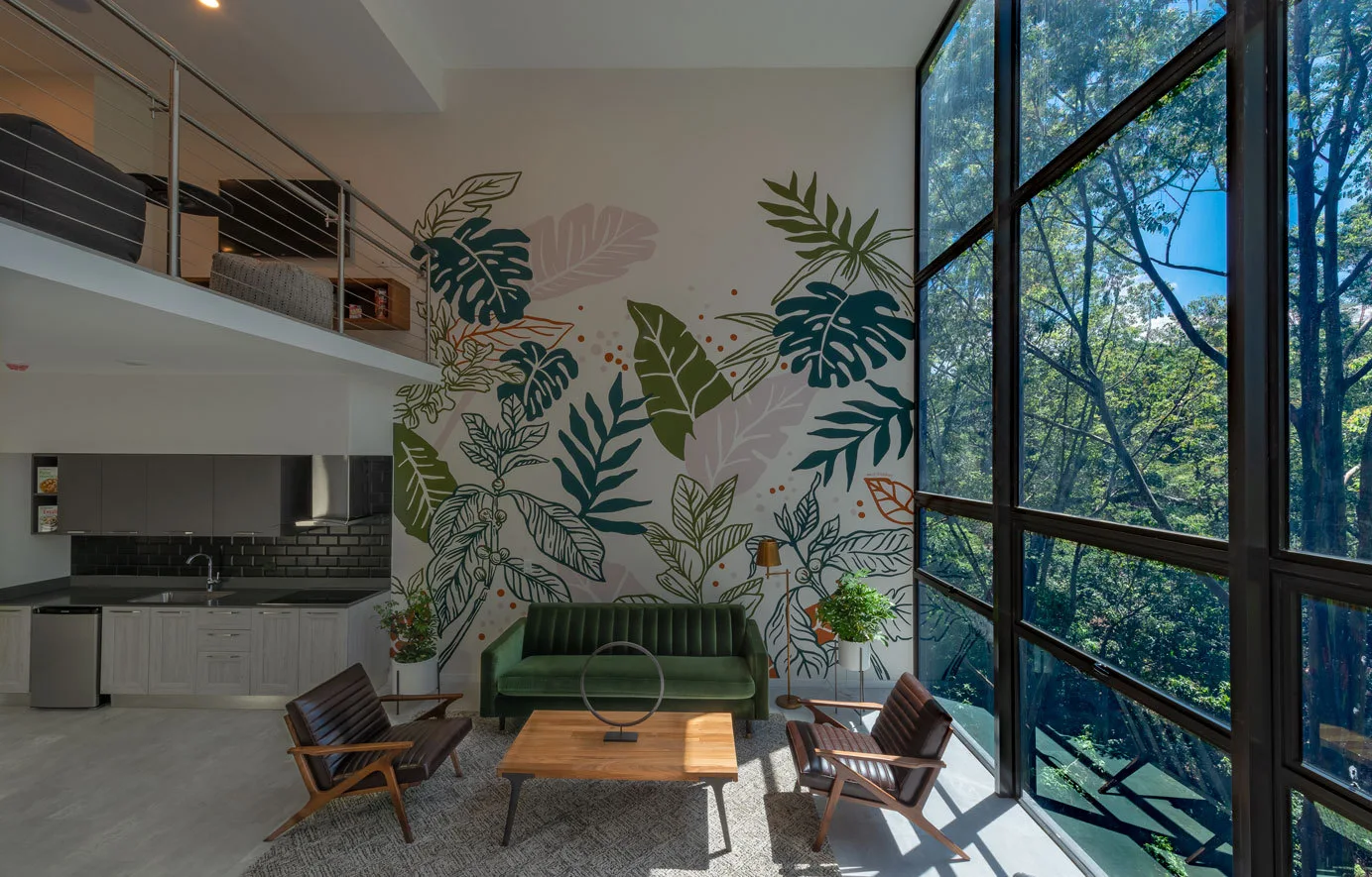 A double-height loft living room featuring a green velvet sofa and brown leather chairs in front of a botanical mural and large multi-paned windows.