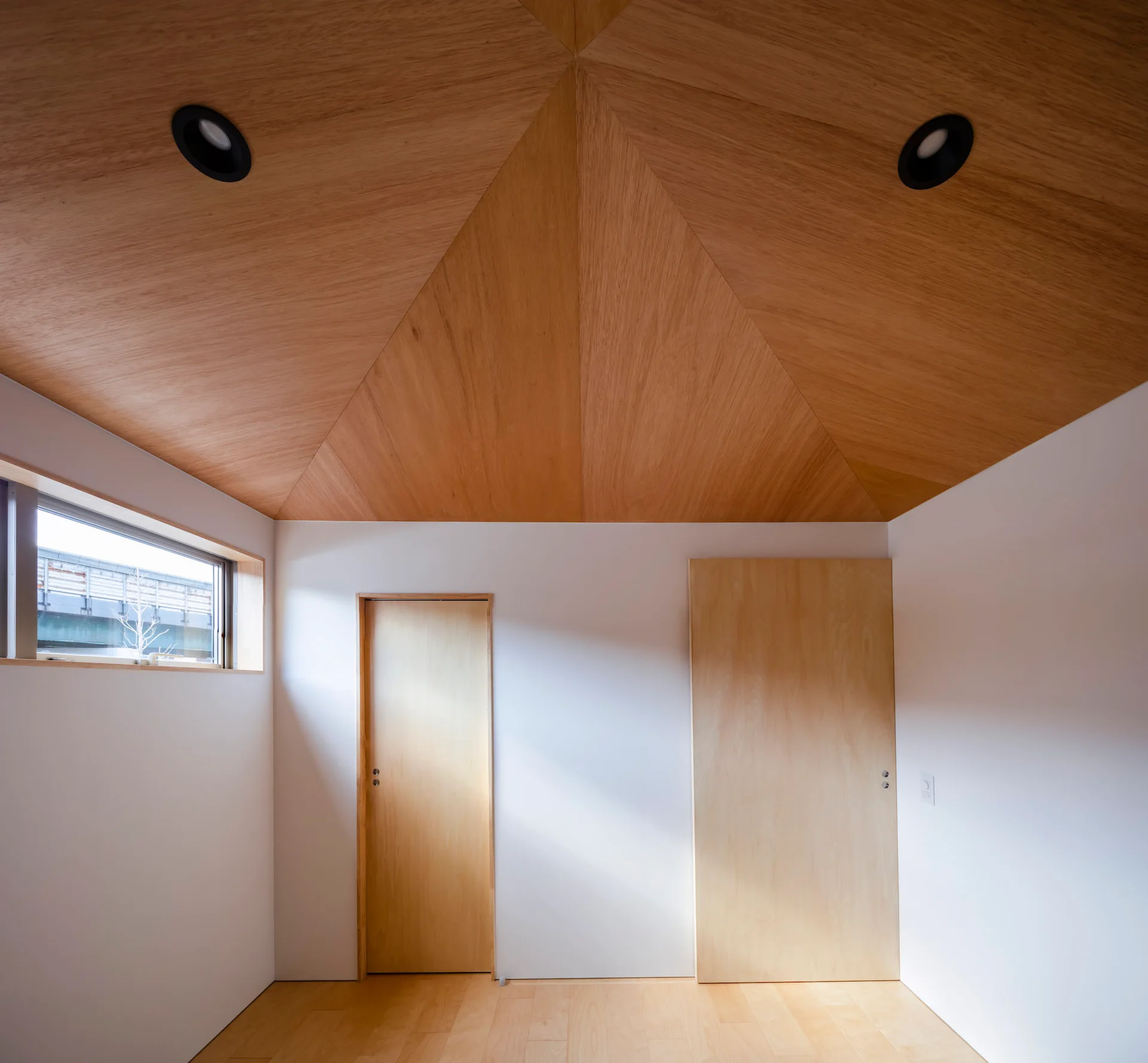 A minimalist room features a geometric vaulted wooden ceiling, matte white walls, two flush light wood doors, and a small horizontal window.
