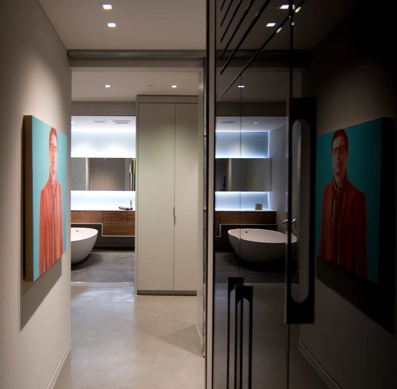 A grey-floored hallway features a colorful portrait painting and leads into a modern bathroom with a freestanding tub and wood vanity.