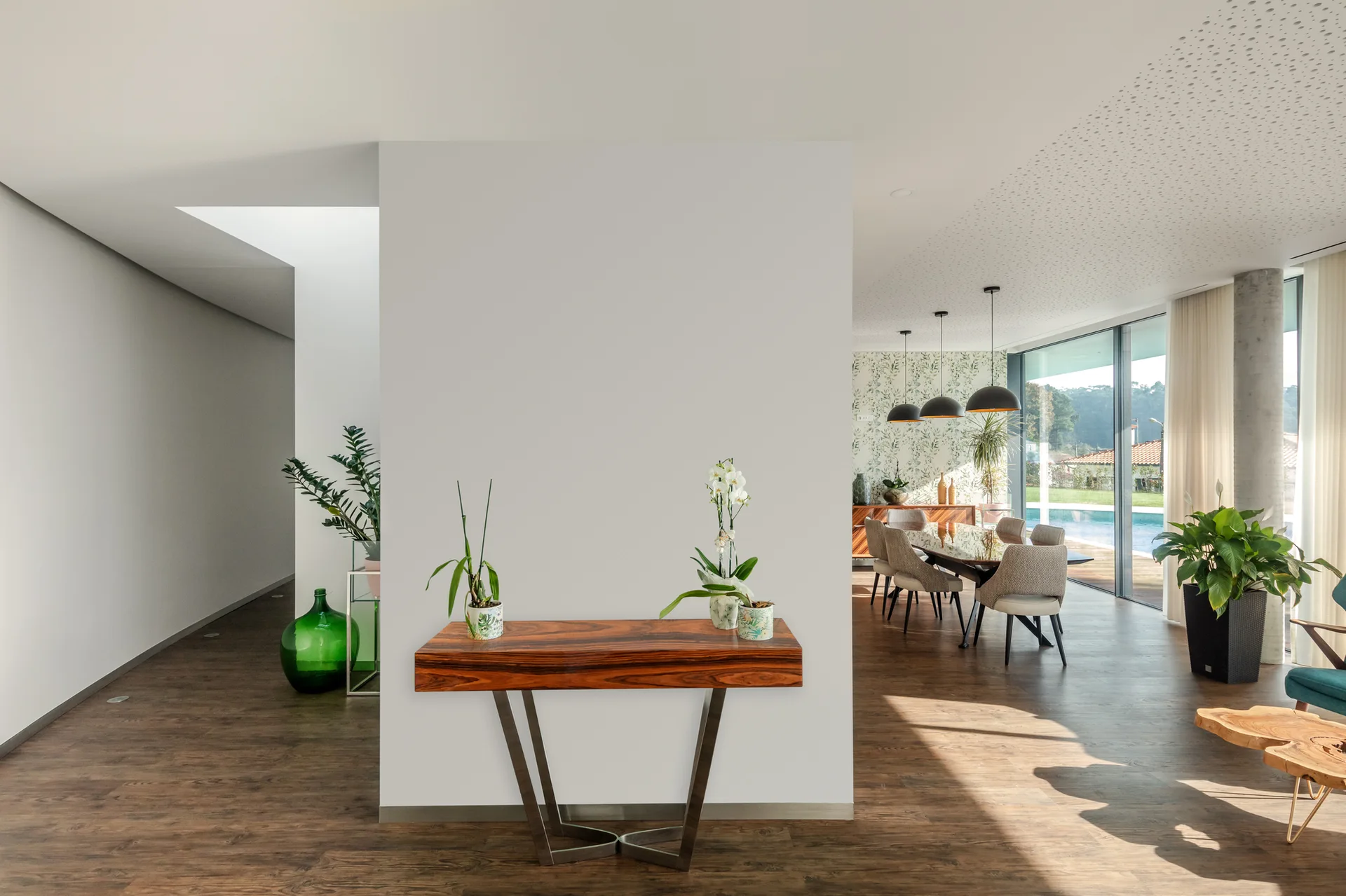 A wooden console table stands against a central white partition wall in an open-plan home, with a dining area and large windows visible behind it.