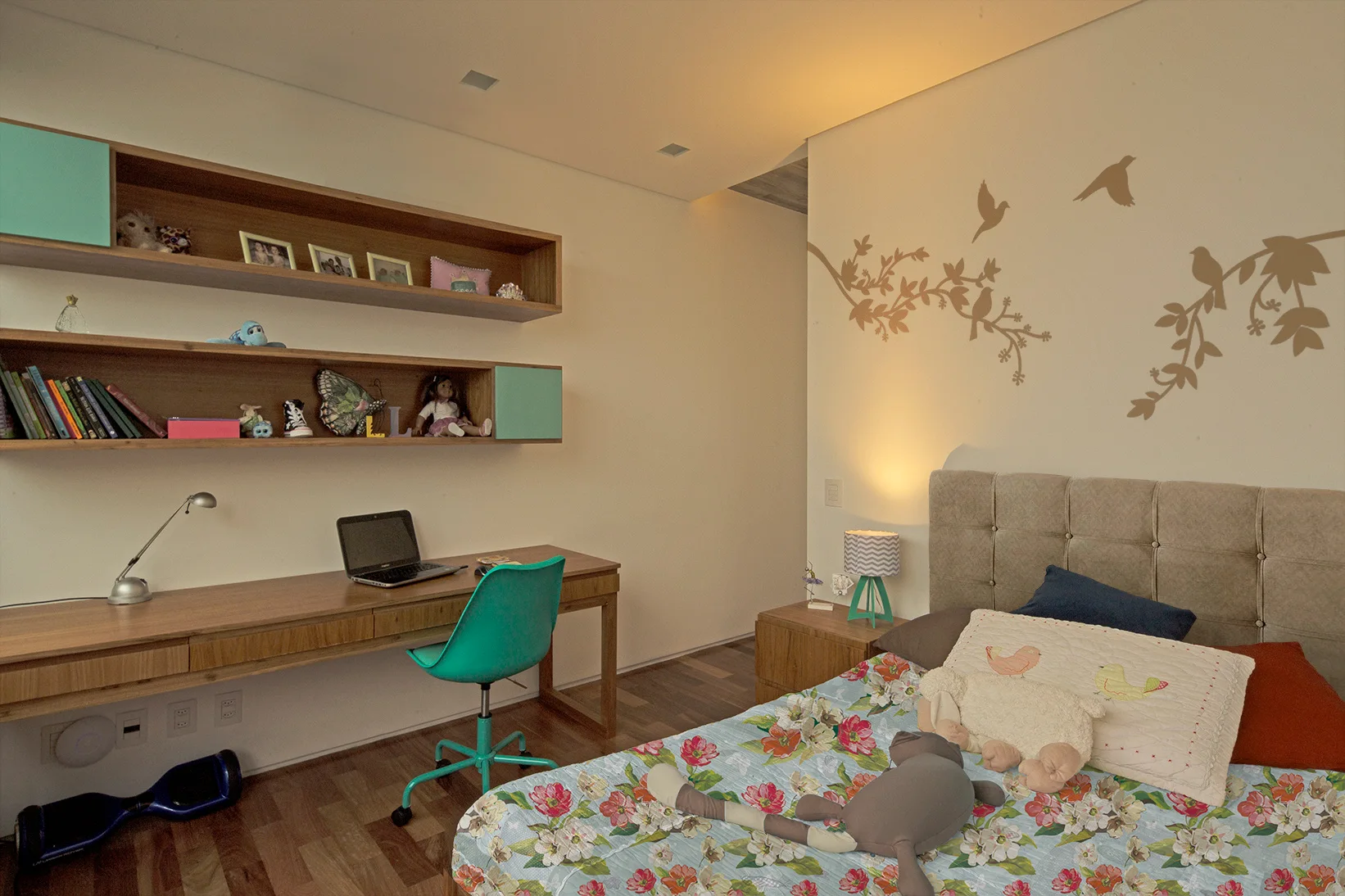 A children's bedroom features a long wooden desk with a teal swivel chair, matching floating shelves on beige walls, and a bed with a tufted headboard.