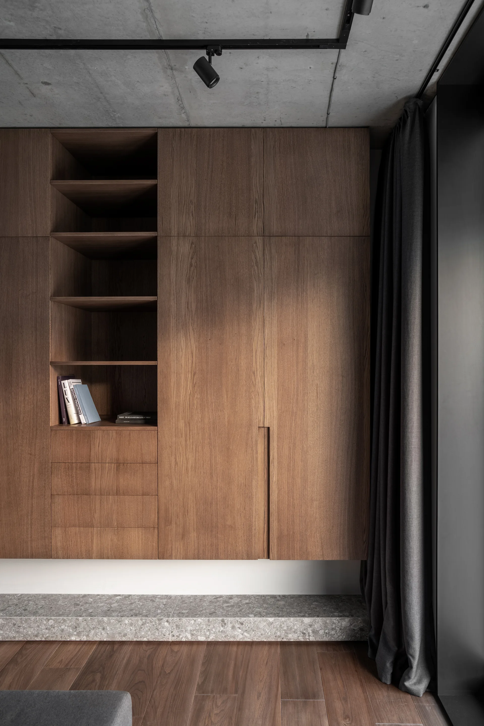 A floating wooden cabinet unit with integrated open shelving sits above a speckled stone platform and wood flooring under an exposed concrete ceiling.