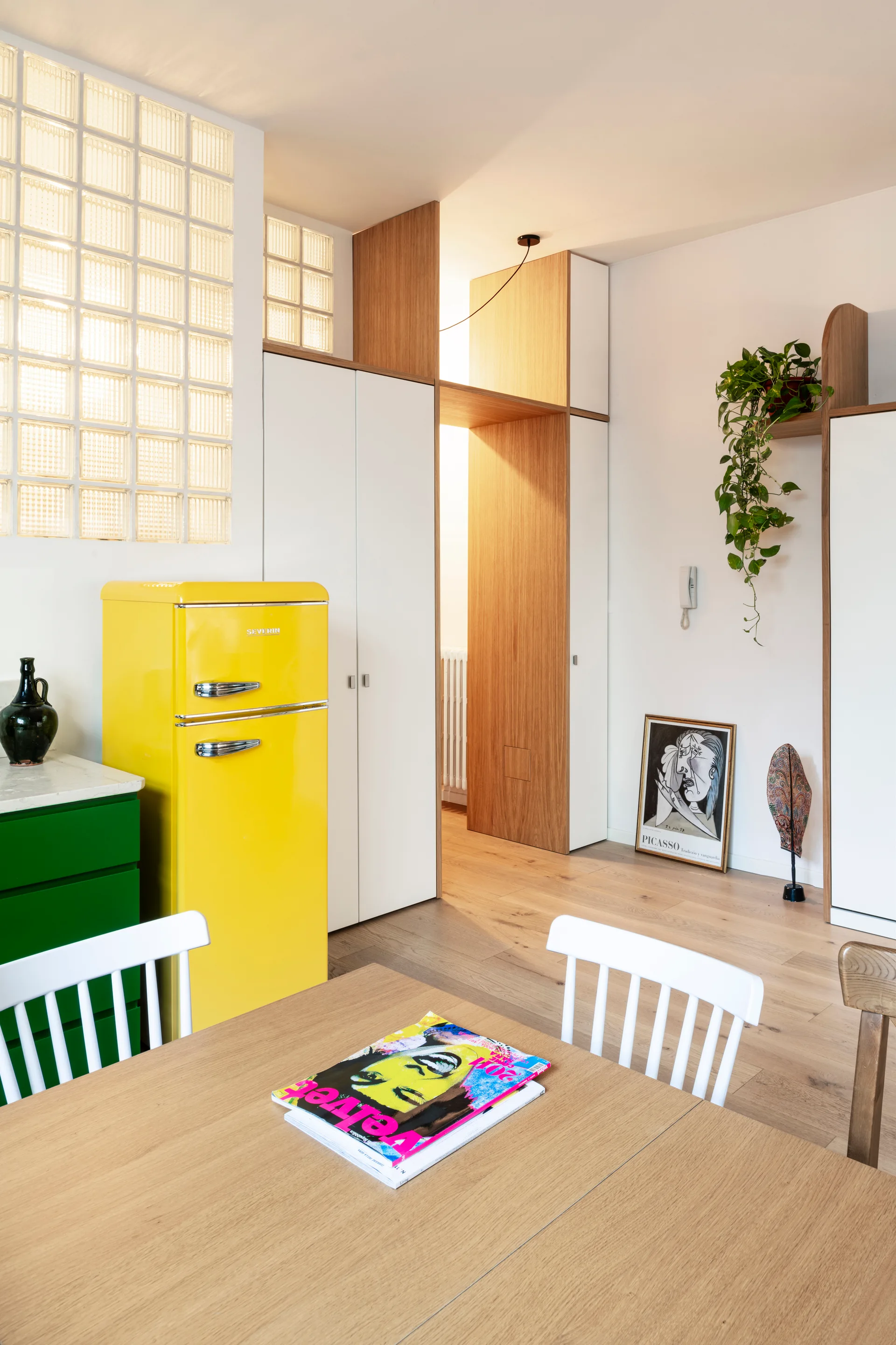 A yellow retro refrigerator stands next to white built-in closets and a glass block partition wall in a light wood floored kitchen.