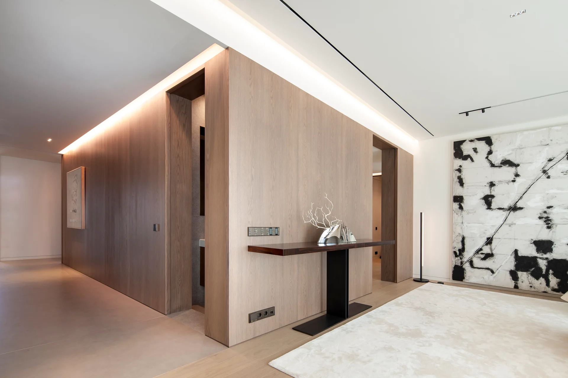 A minimalist hallway features wood paneling with warm cove lighting, a slim console table, and a large black and white painting.
