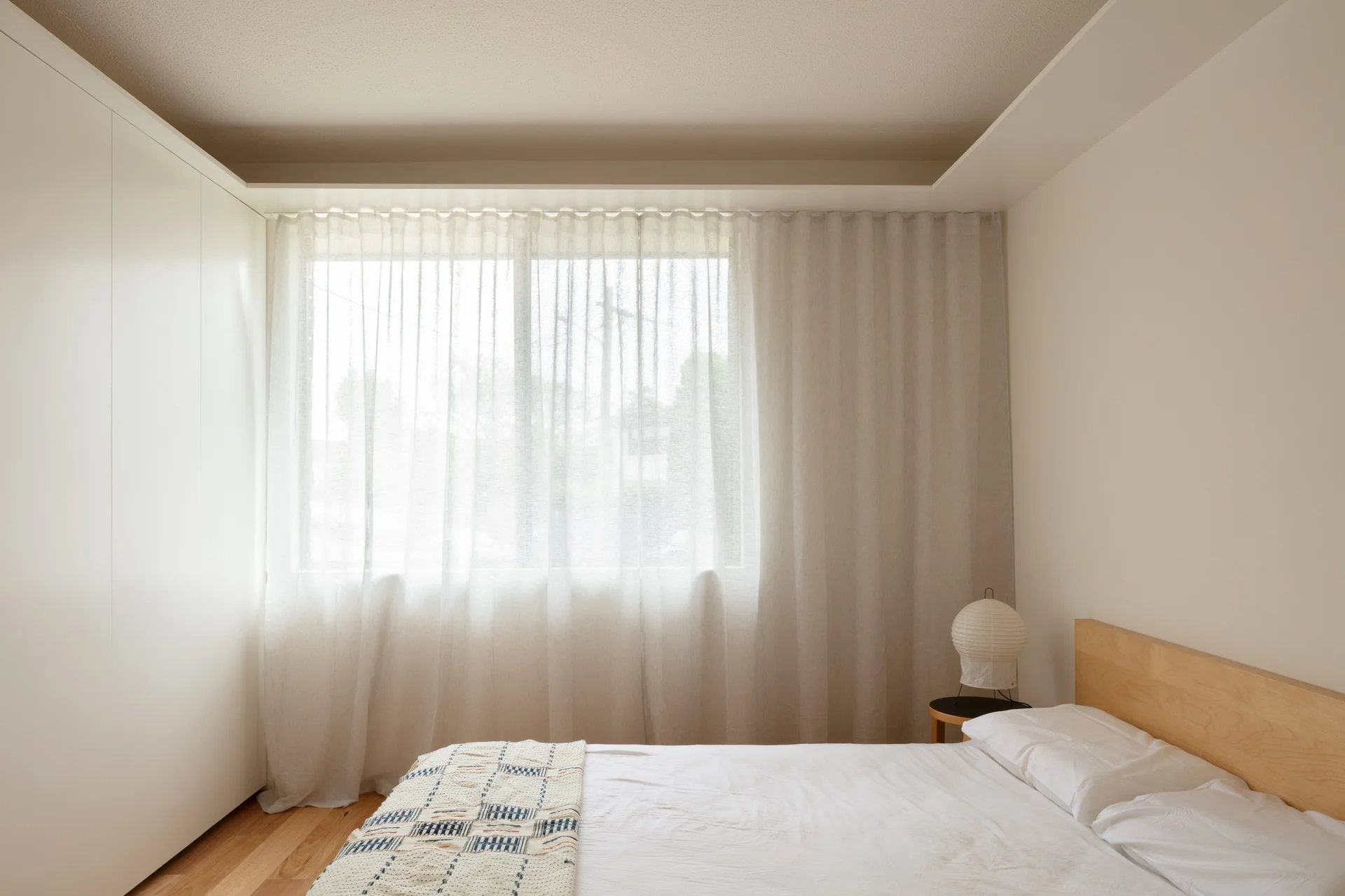 A minimalist bedroom features a light wood headboard, a white built-in wardrobe, and large windows softened by translucent sheer curtains.