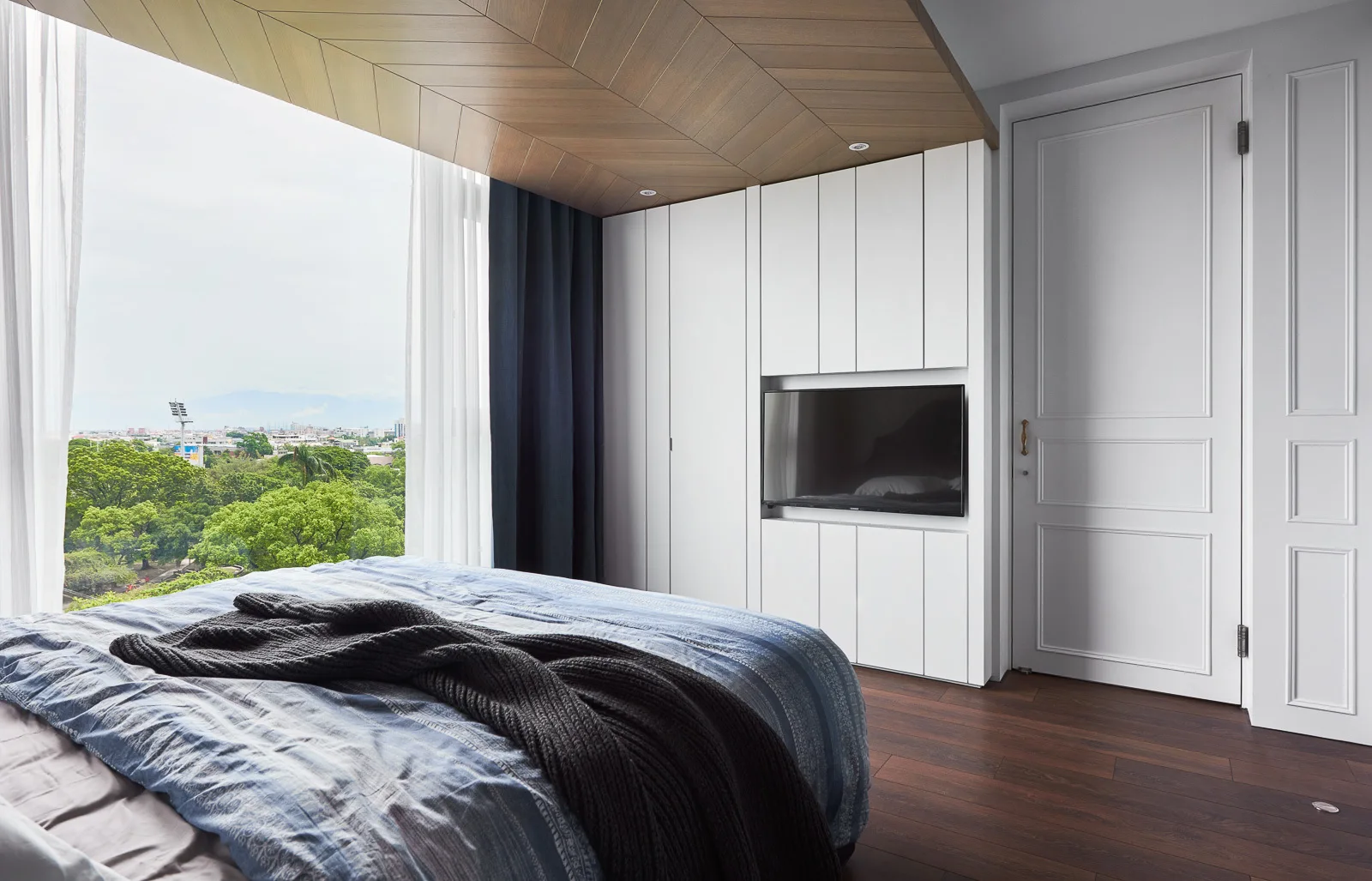 A modern bedroom features a bed with blue bedding, a chevron-patterned wood ceiling, built-in white cabinets with a recessed television, and large windows.