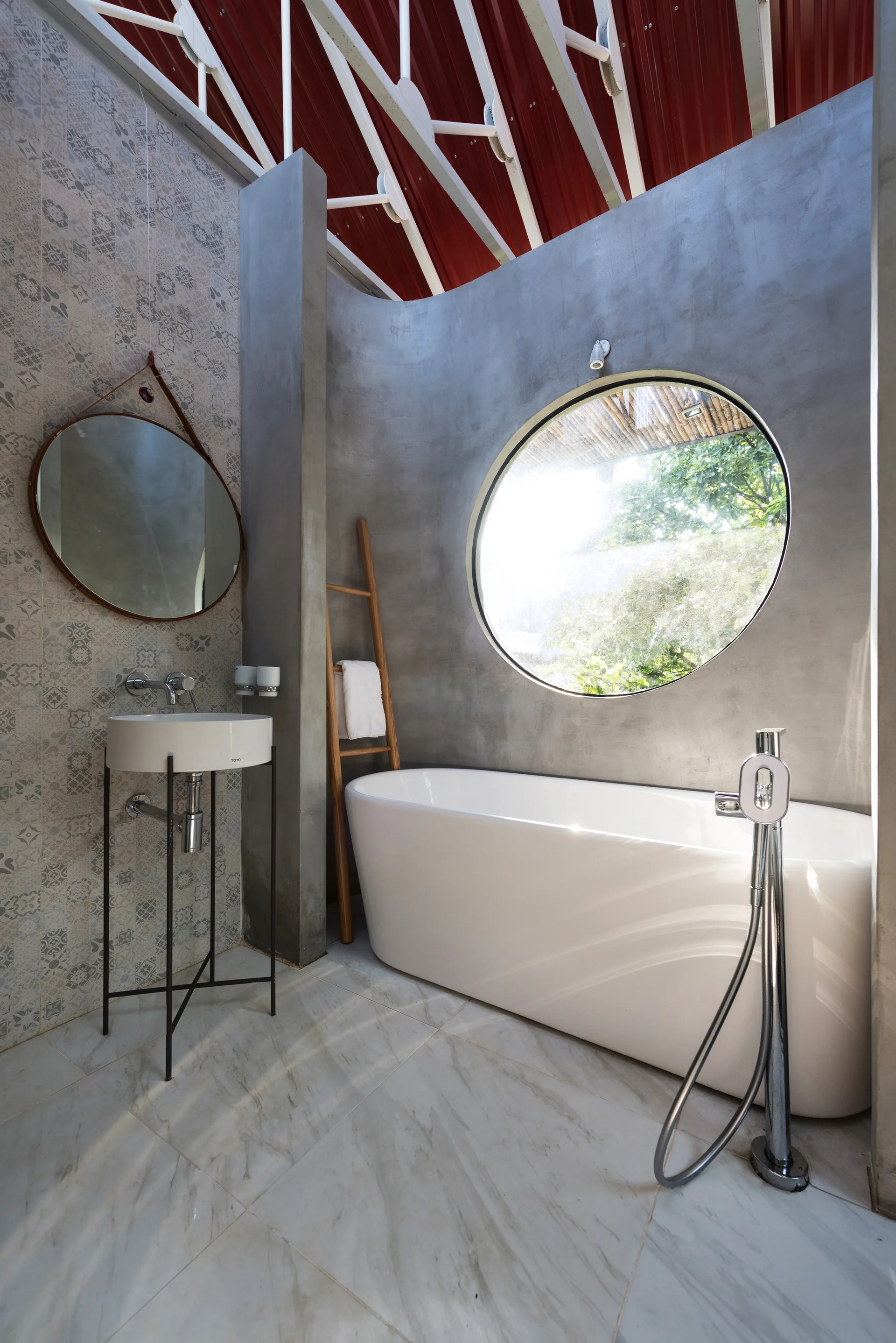 A modern bathroom featuring a white freestanding tub, a circular window in a concrete wall, and patterned grey tiles under a red metal roof.