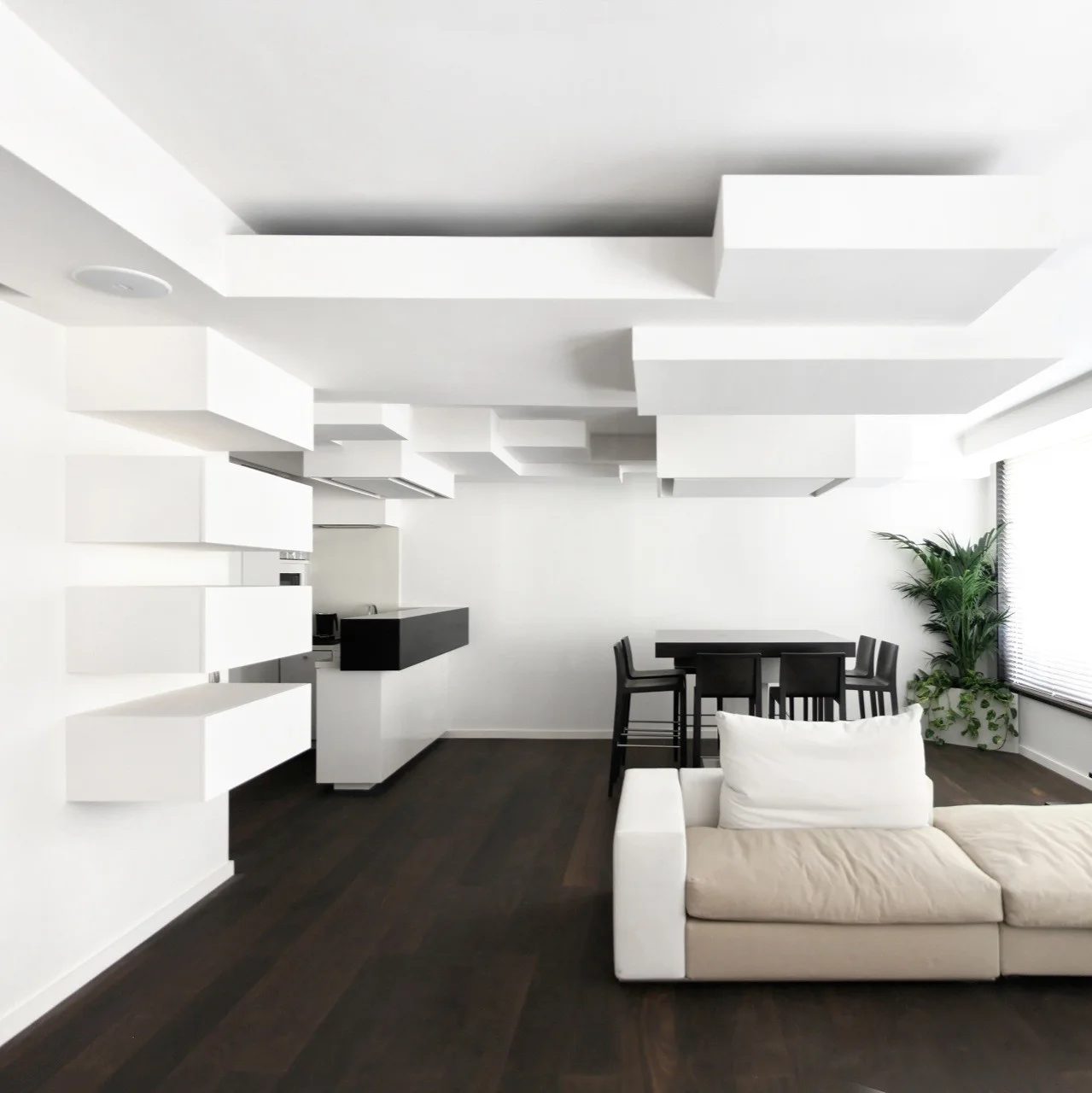 A minimalist open-plan living room with sculptural white ceiling blocks, stacked floating shelves, a cream sofa, and dark wood flooring.