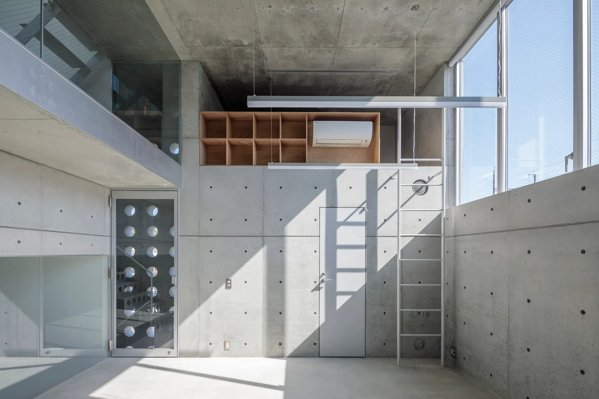A minimalist room features exposed concrete walls, a tall metal ladder, large windows, and a wooden grid shelving unit.