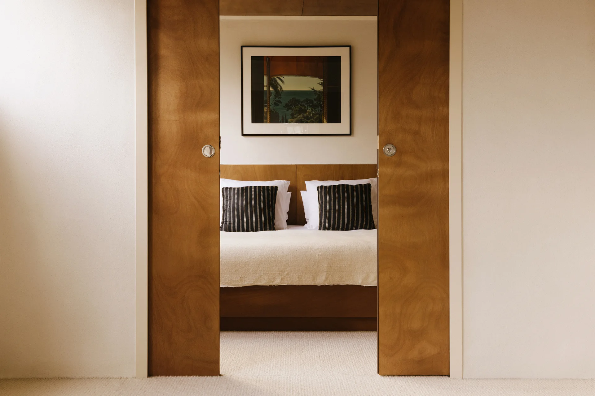 A view through open wooden pocket doors into a bedroom with a wood-paneled headboard, white bedding, striped pillows, and a framed art print.