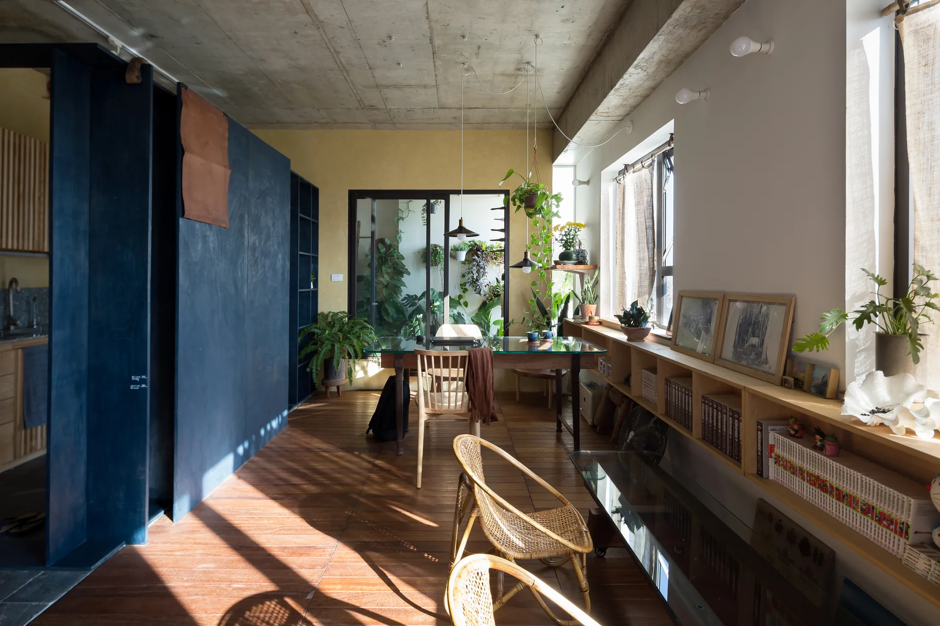 An industrial living room features a raw concrete ceiling, a low wooden bookshelf under windows, a dark blue partition wall, and rattan chairs.
