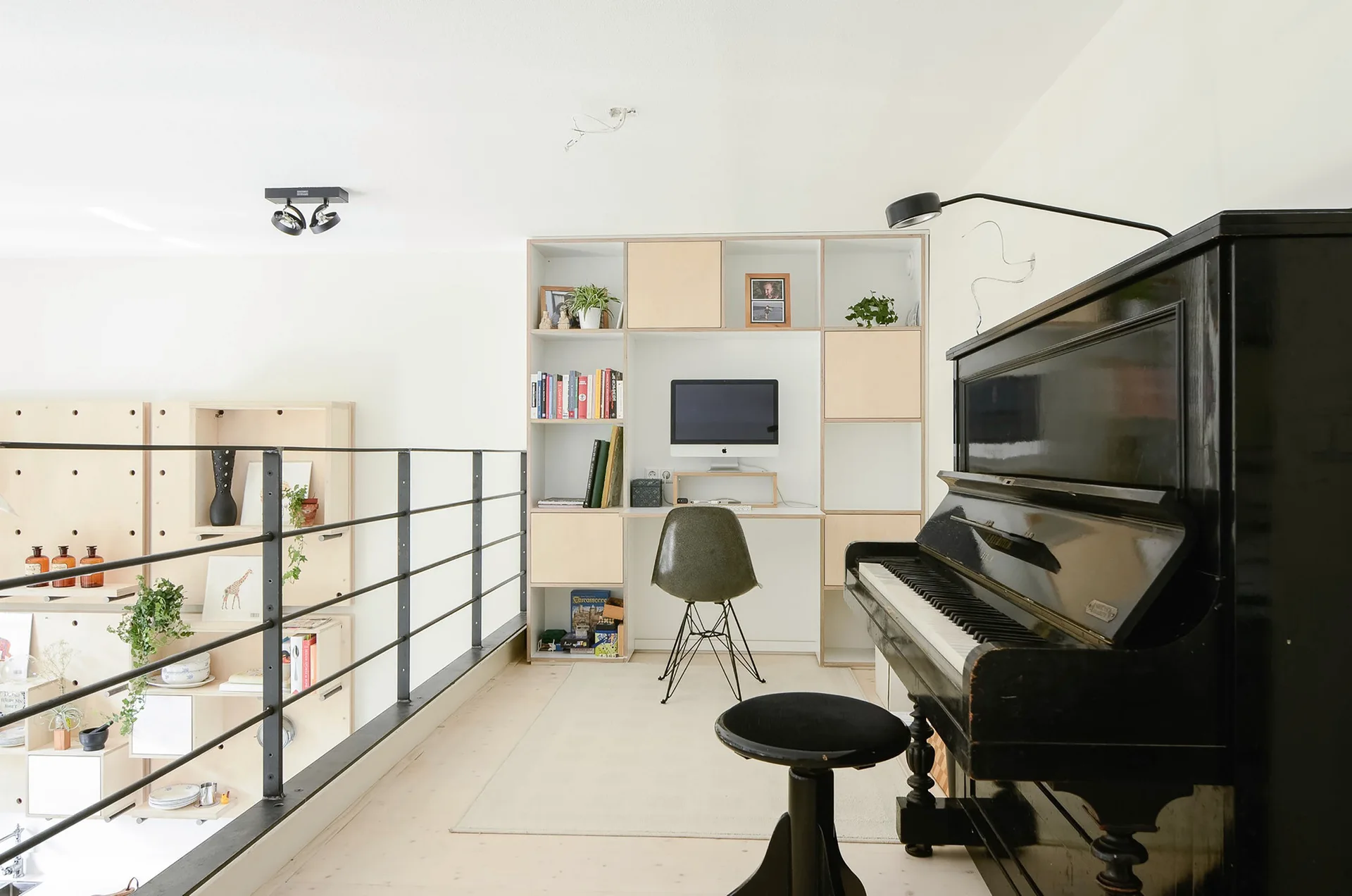 A mezzanine home office workspace features a built-in light wood shelving unit with a desk, a black upright piano, and a black metal railing.