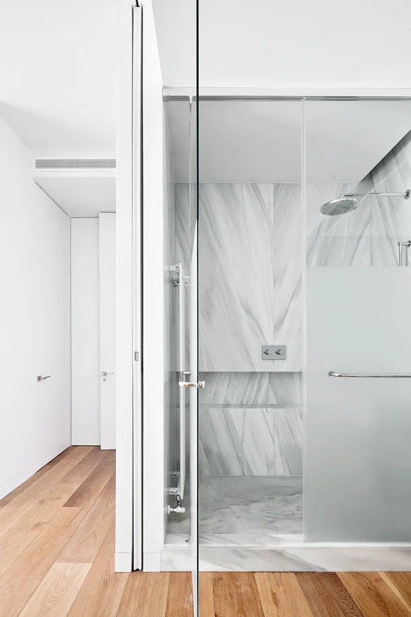 A glass-enclosed walk-in shower lined with veined white marble stands adjacent to a hallway with light hardwood floors.