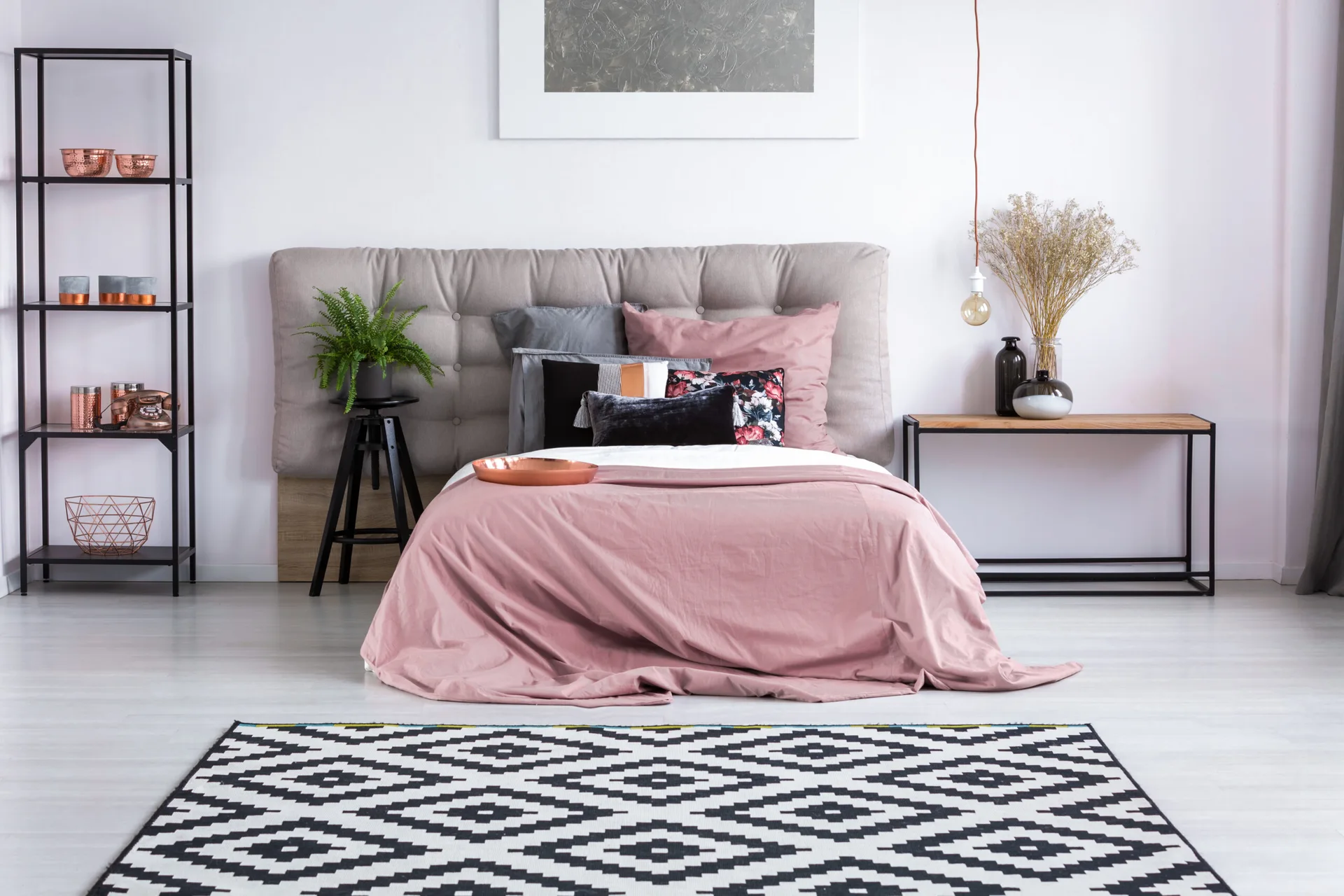 A modern bedroom features a bed with pink bedding and a grey tufted headboard, set above a black and white geometric area rug.
