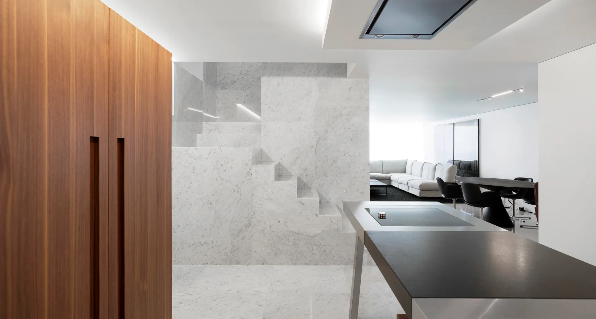 A contemporary open-concept space featuring a solid white marble staircase, a warm wood cabinet, and a dark kitchen island.