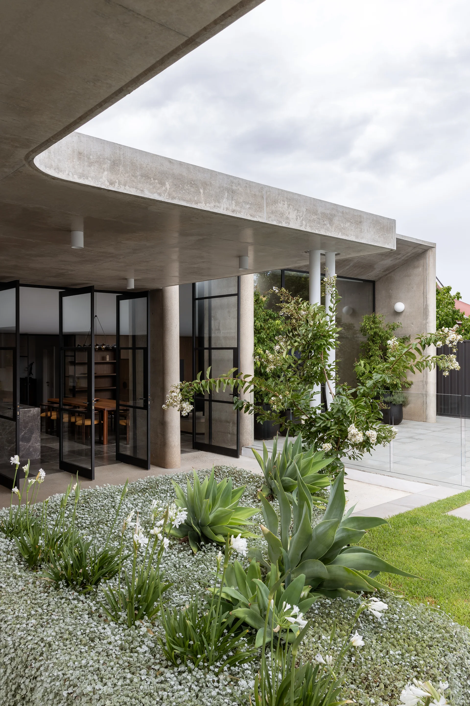 A modern concrete patio features an overhanging roof, black metal-framed glass doors, and a landscaped garden bed with large green agave plants.