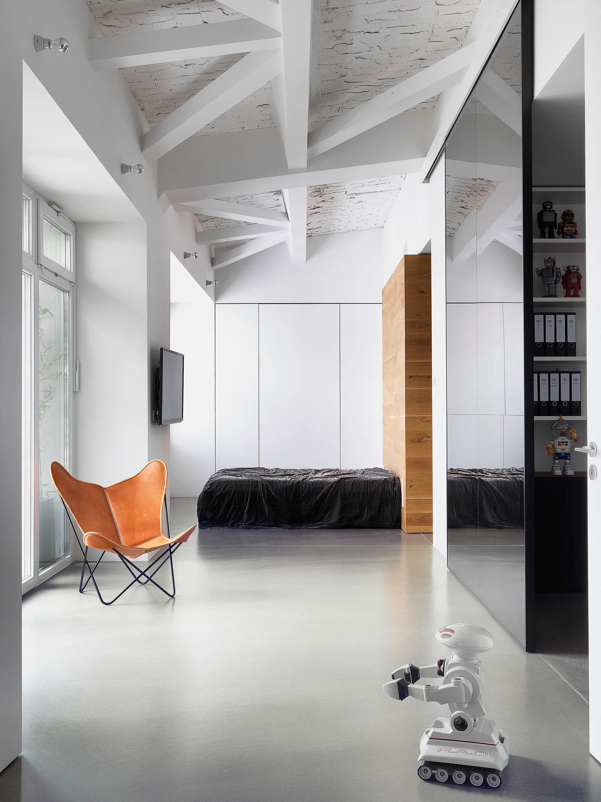 A minimalist bedroom featuring a tan leather butterfly chair, a low platform bed, exposed white ceiling beams, and a mirrored sliding wardrobe.