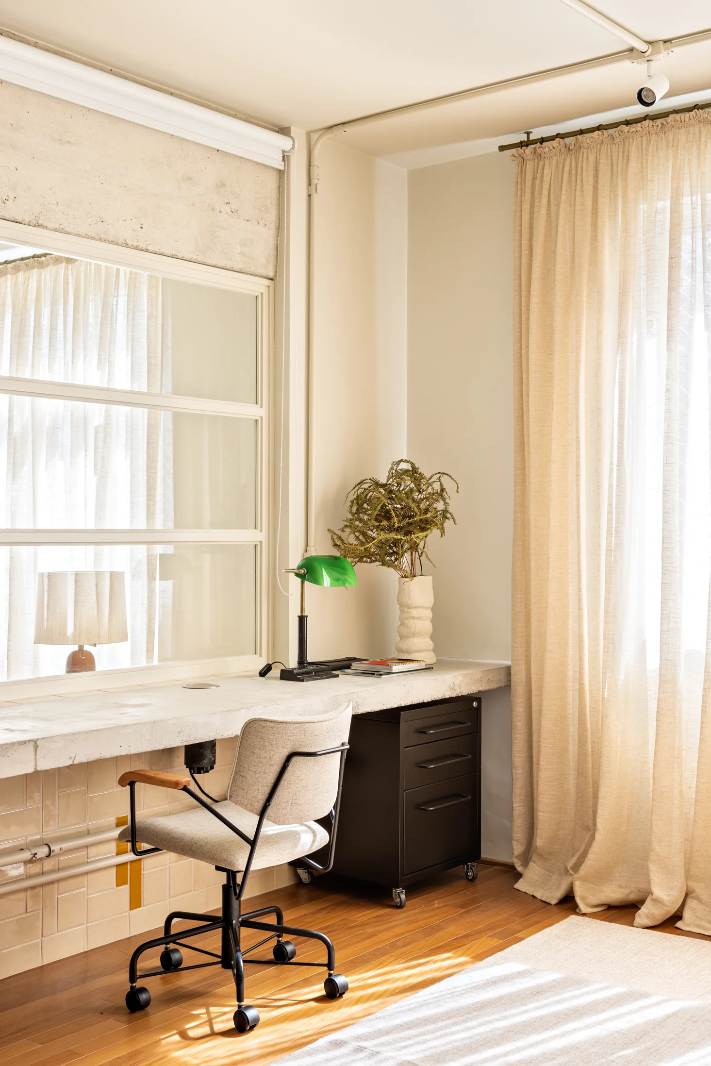 A minimalist home office features a concrete desk slab, a neutral fabric desk chair, a green task lamp, and linen curtains next to a window.