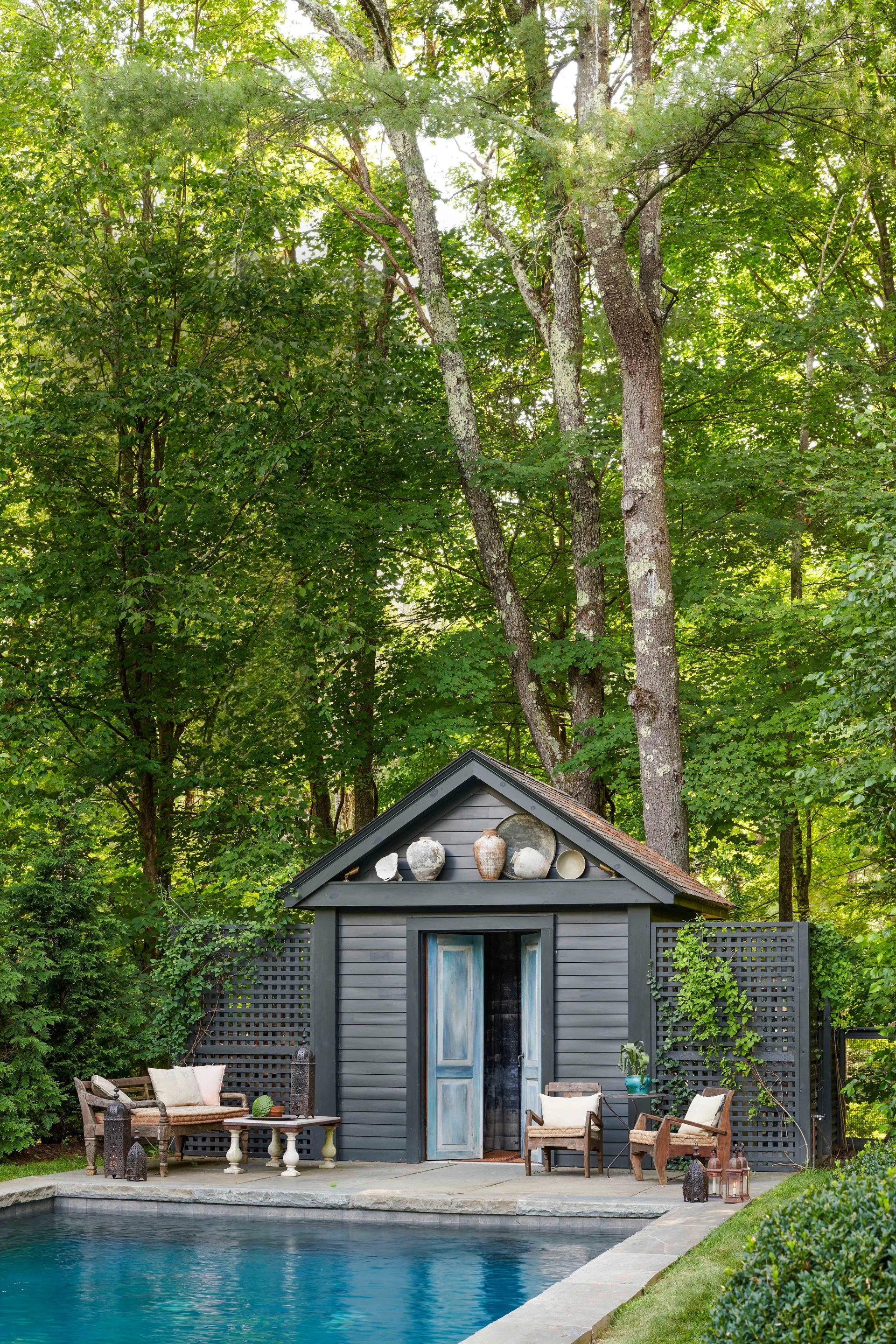 A charcoal gray pool house with weathered blue doors sits behind a stone pool deck with wooden patio furniture under tall green trees.